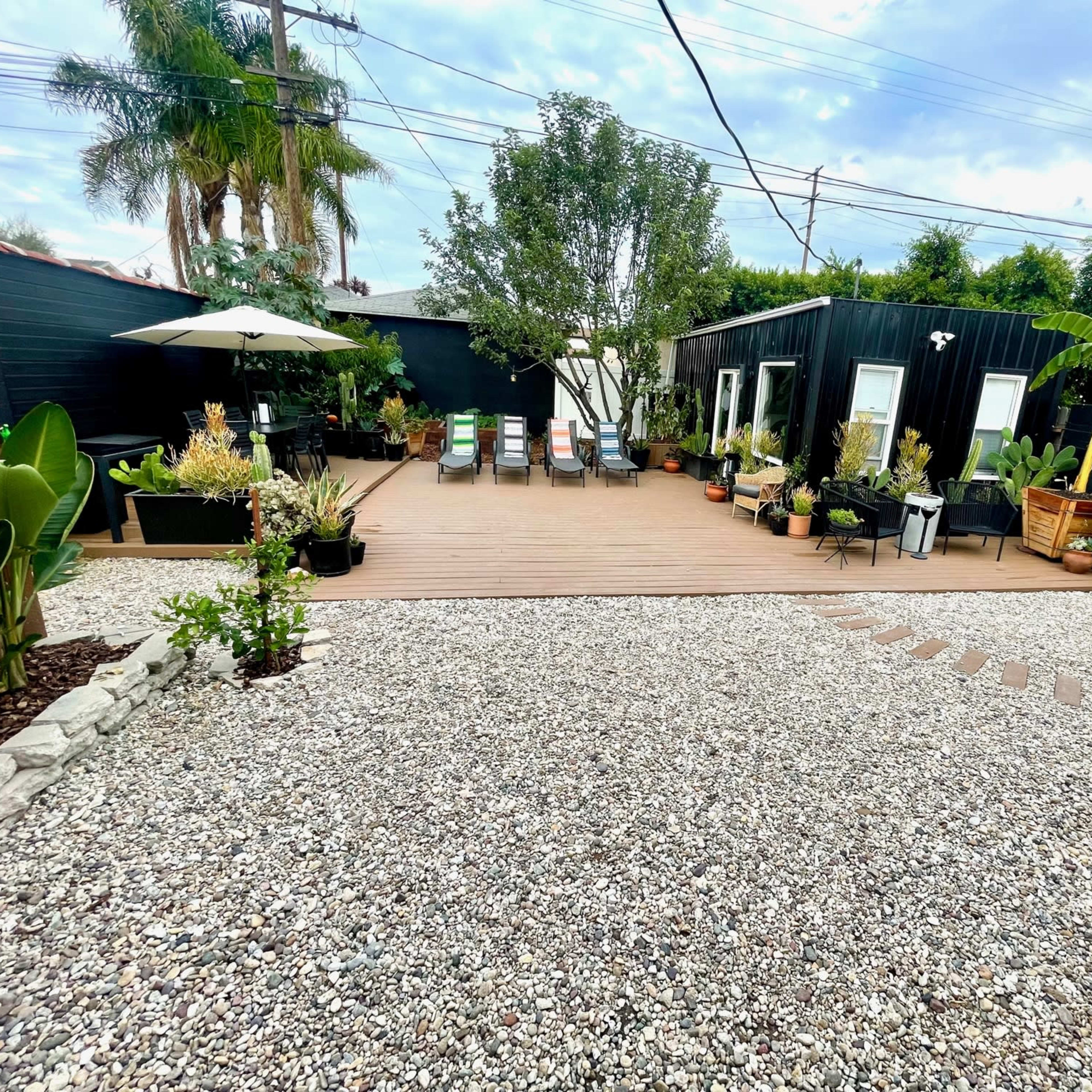 A backyard featuring a wooden deck surrounded by potted plants, a gravel path, and several lounge chairs.