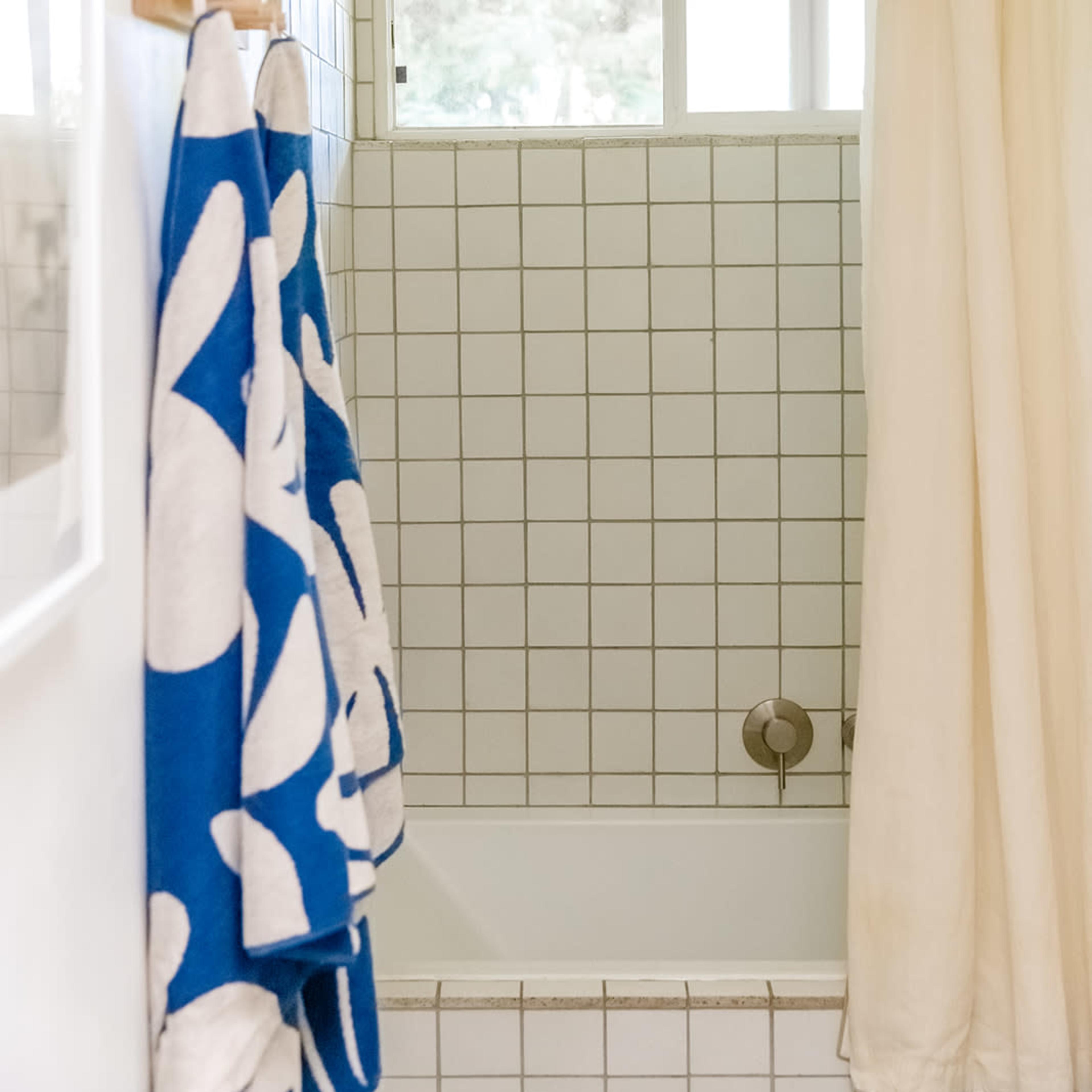 A bathroom with a tiled shower area, a bathtub, a light-colored shower curtain, and two towels hanging on a hook.