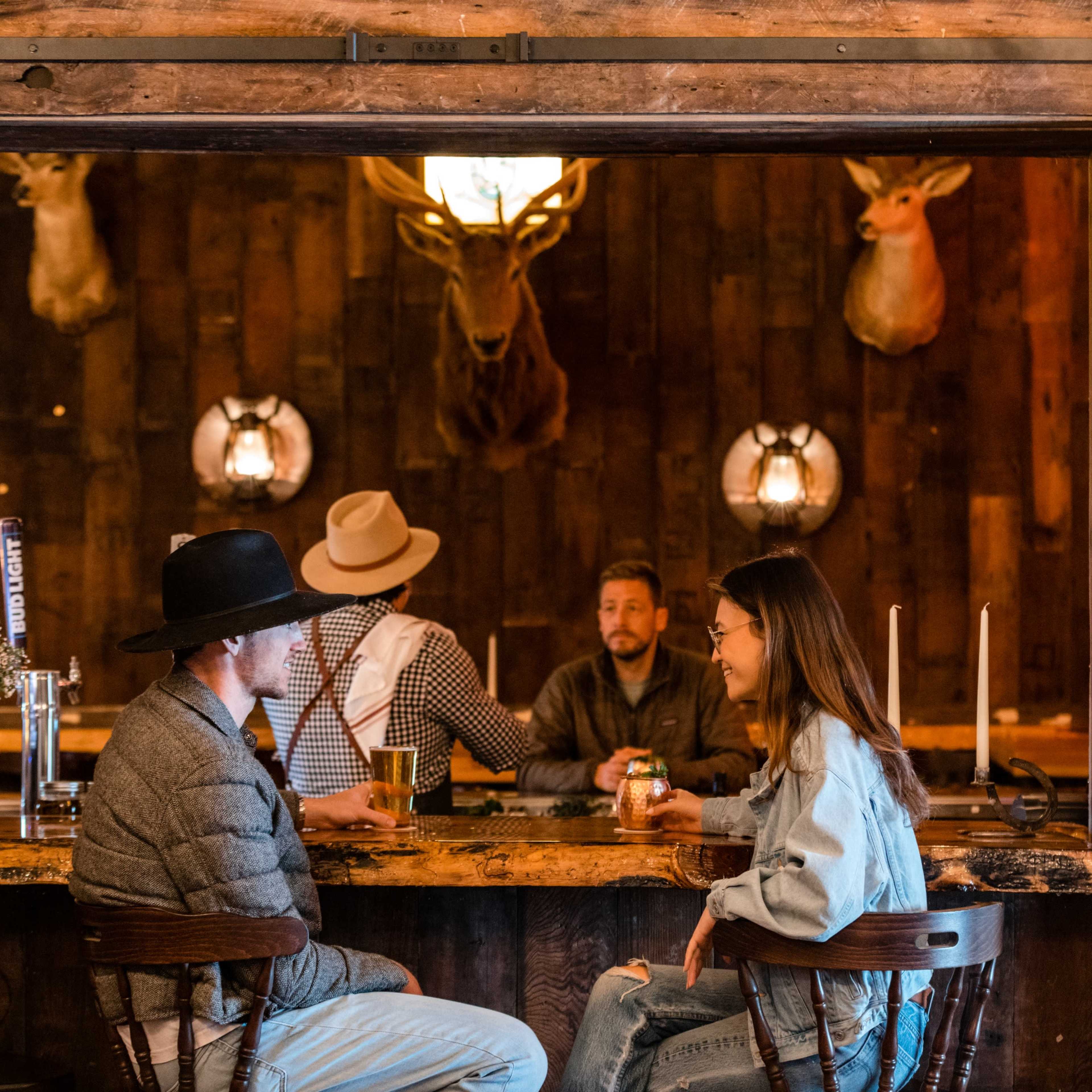 A bar scene features two patrons conversing at the counter while a bartender and another customer are visible in the background, with mounted deer heads on the wall.