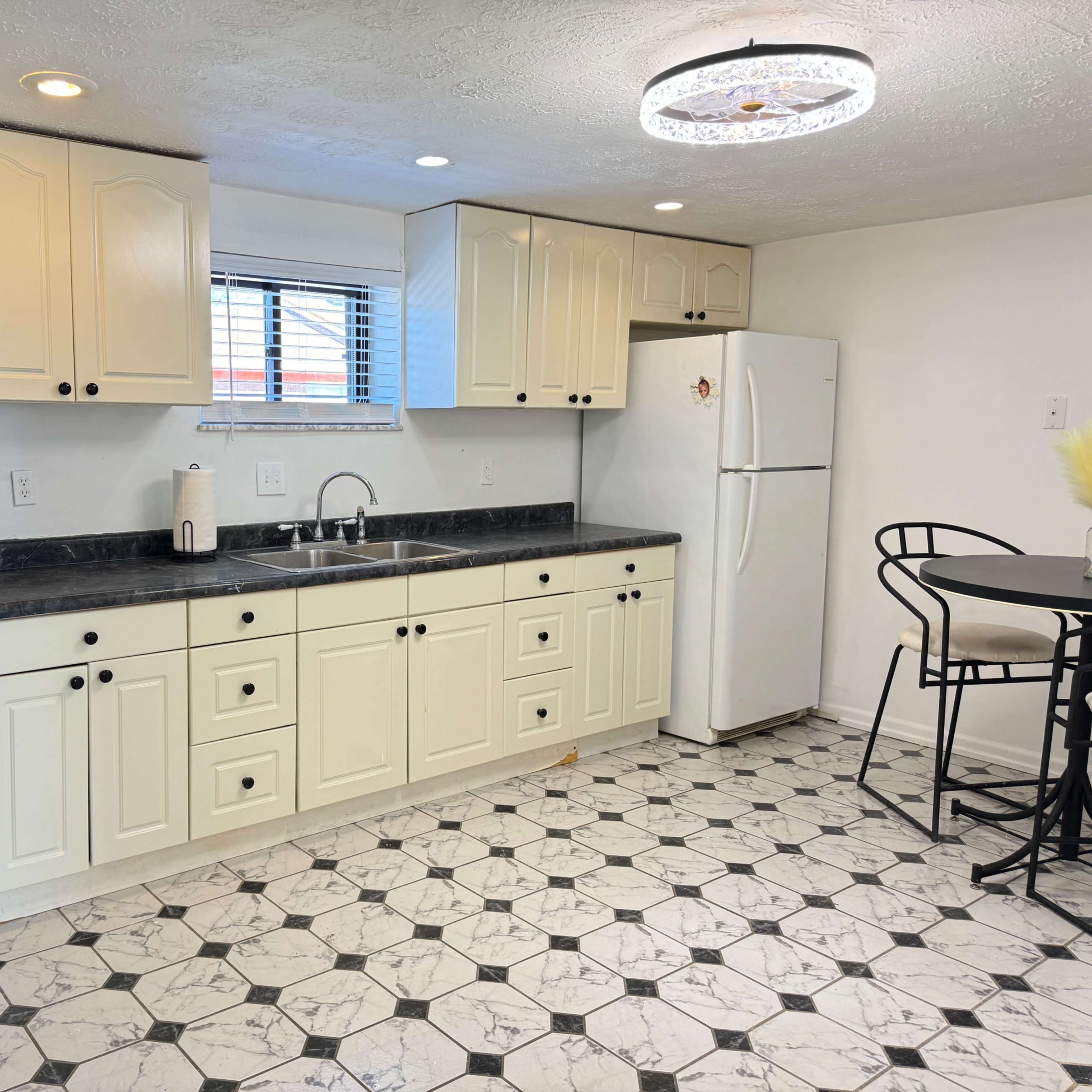 A kitchen with light-colored cabinets, a black countertop, a white refrigerator, an oven, and a small dining table with black chairs.