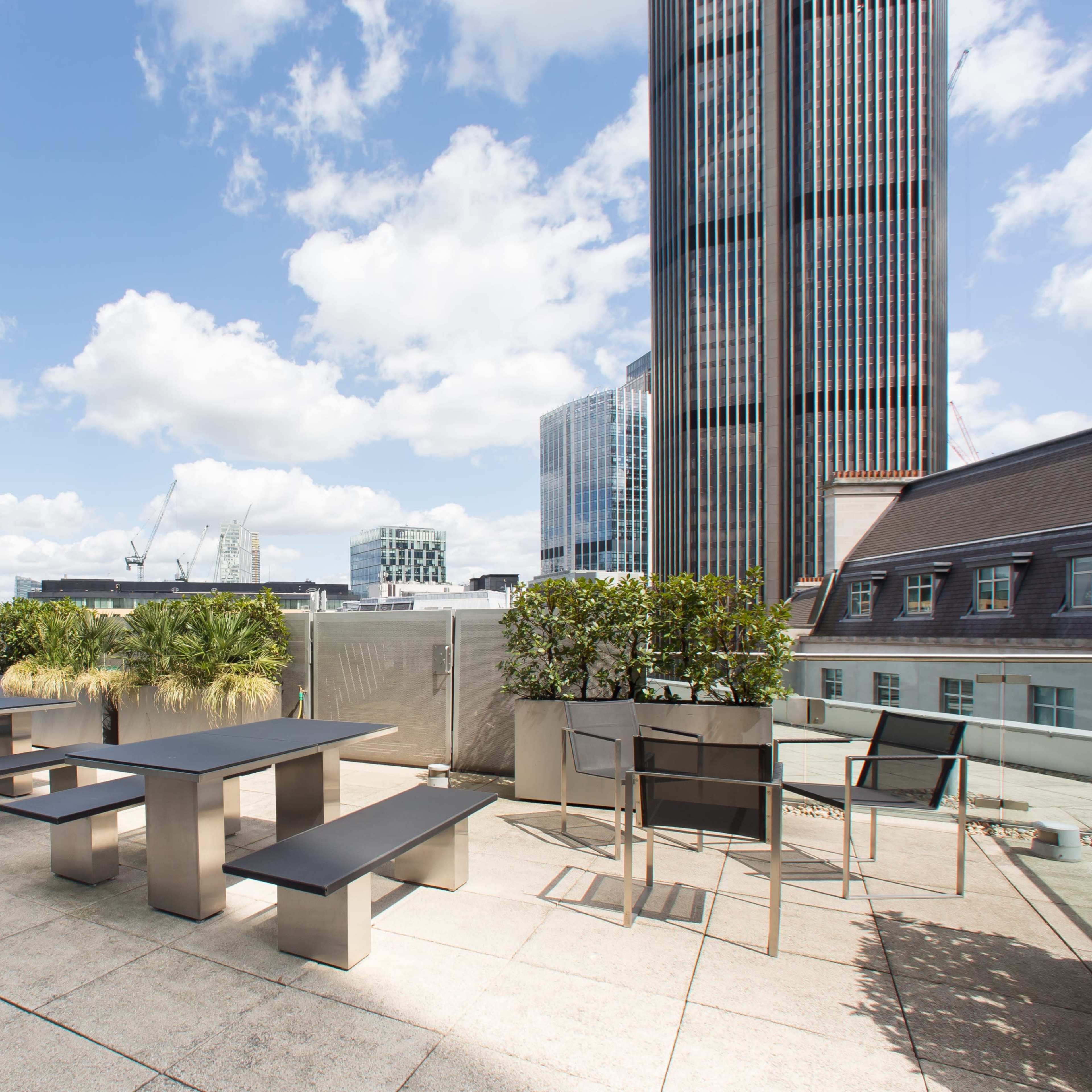 A rooftop terrace with modern tables and benches surrounded by potted plants, set against a backdrop of tall buildings and a blue sky.
