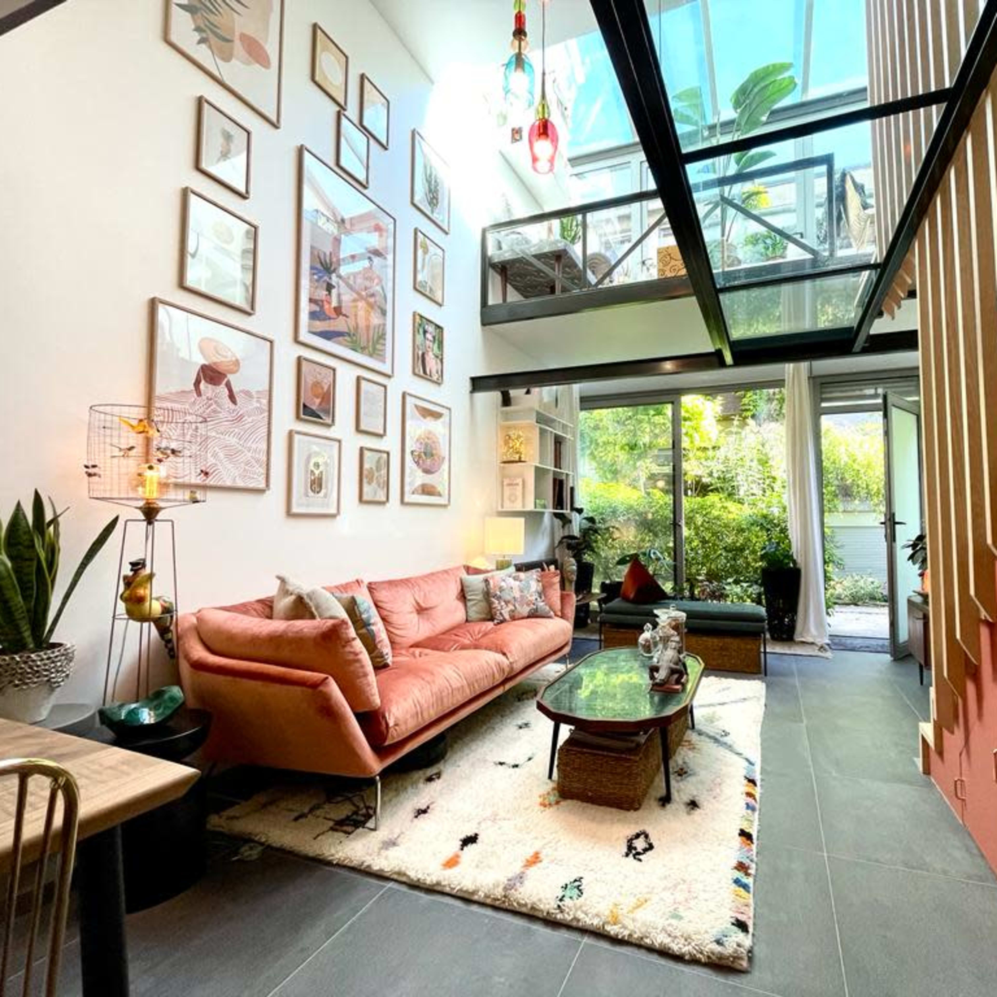 A modern living space featuring a pink sofa, a coffee table, and a glass-walled balcony overlooking a garden, with various framed artworks adorning the walls.