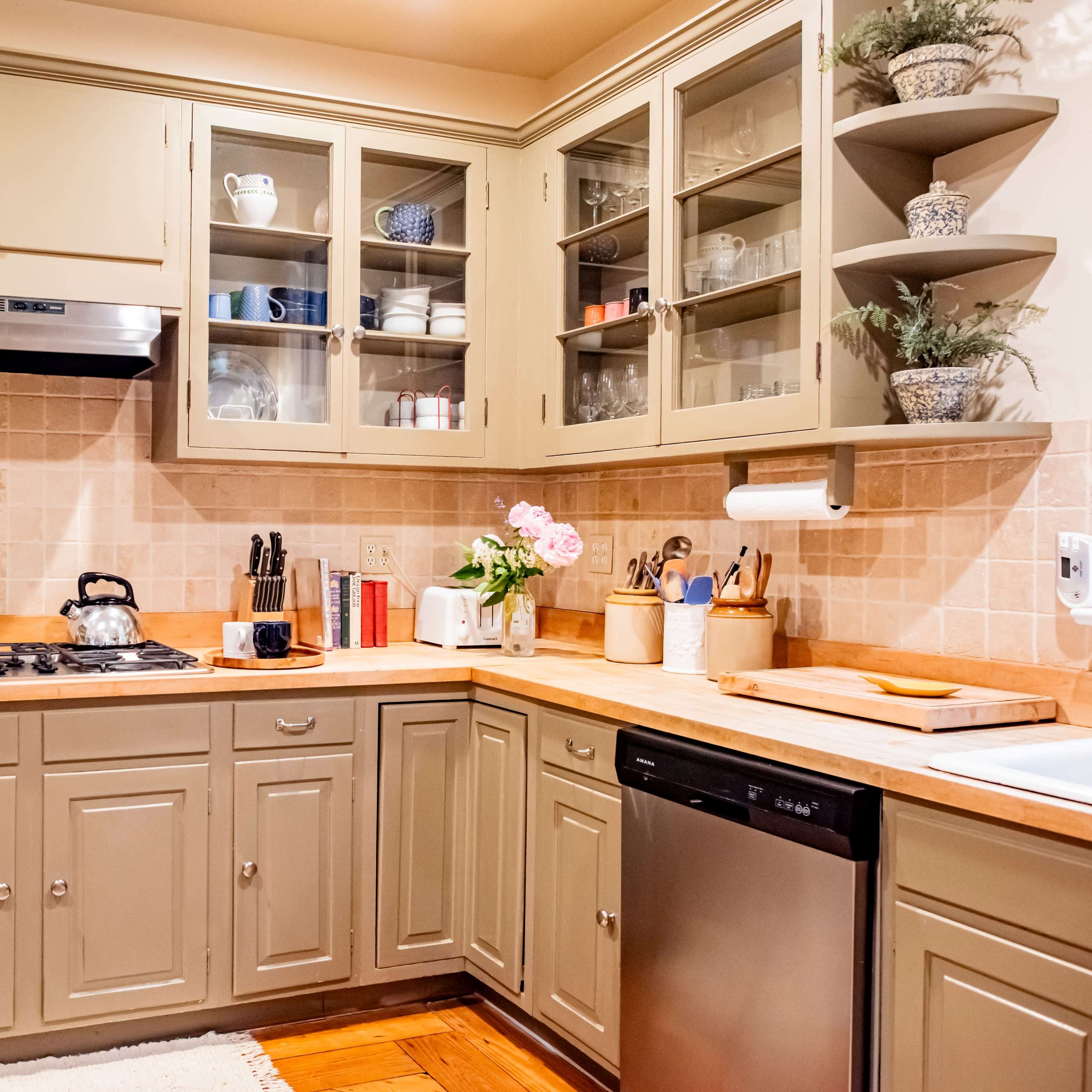 A well-organized kitchen with beige cabinetry, a gas stove, a stainless steel dishwasher, and various kitchenware displayed on open shelves.