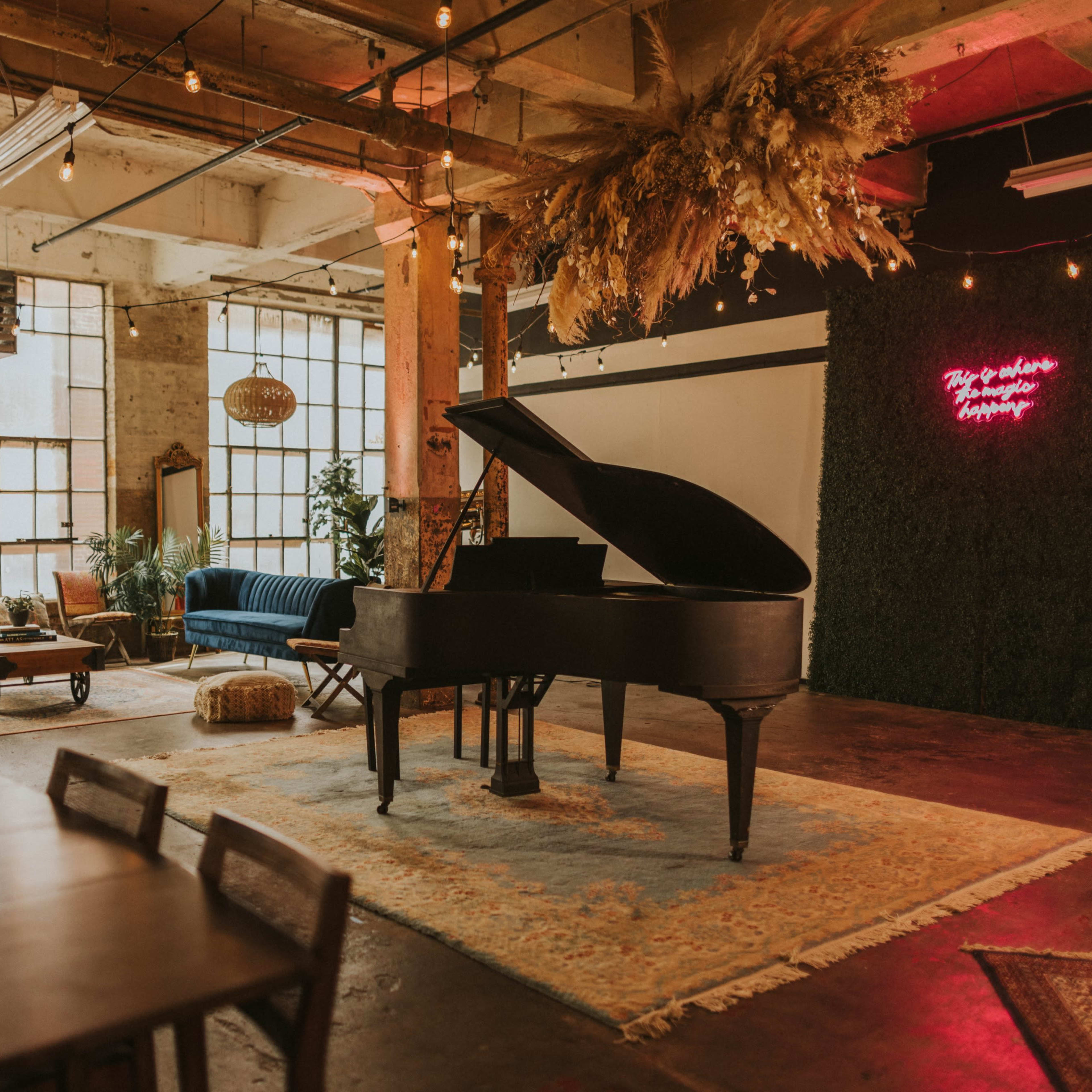 A grand piano sits on a rug in a spacious, well-lit room with large windows, surrounded by modern seating and decorative plants.