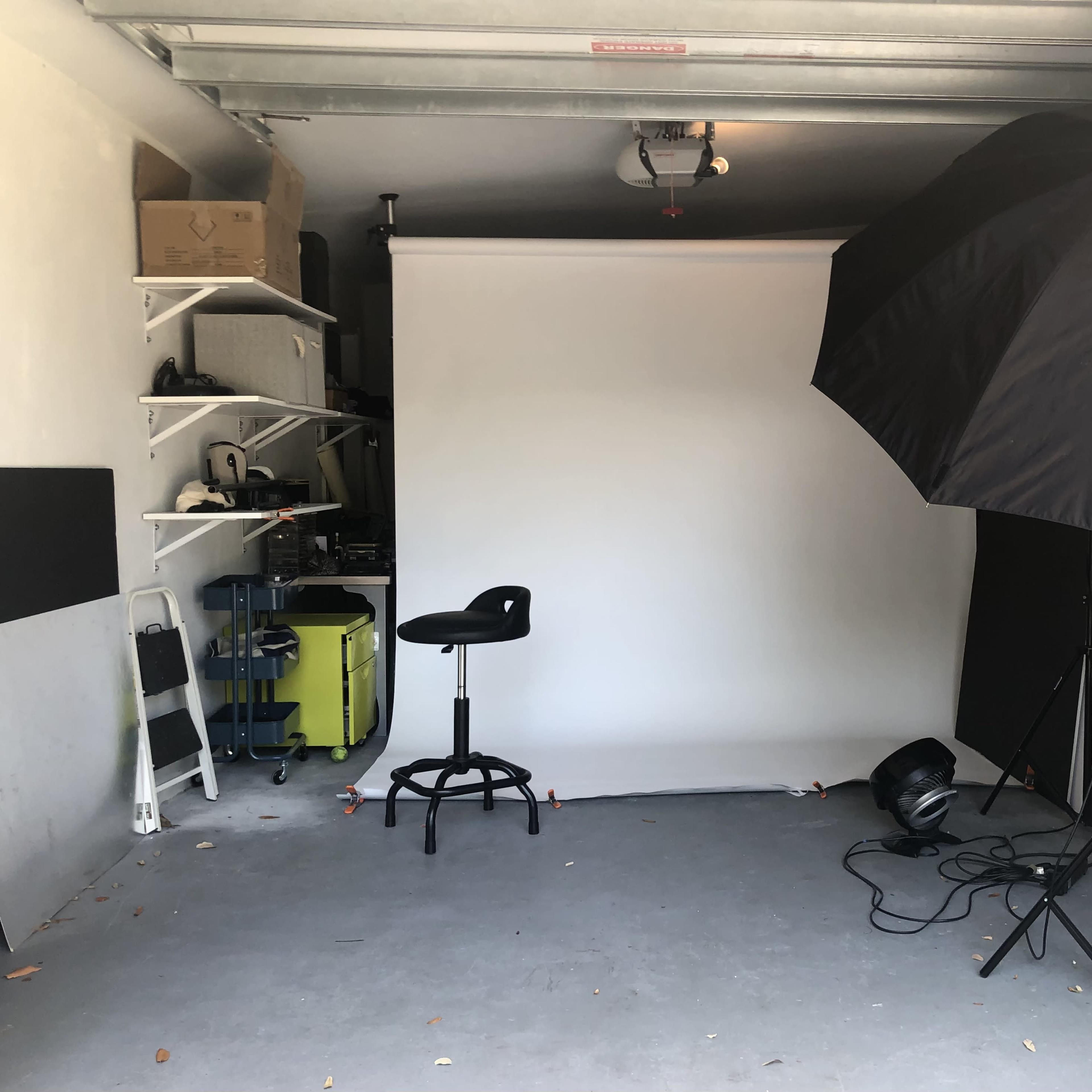 A garage setup with a white backdrop, a black umbrella light, a stool, and various equipment on shelves.