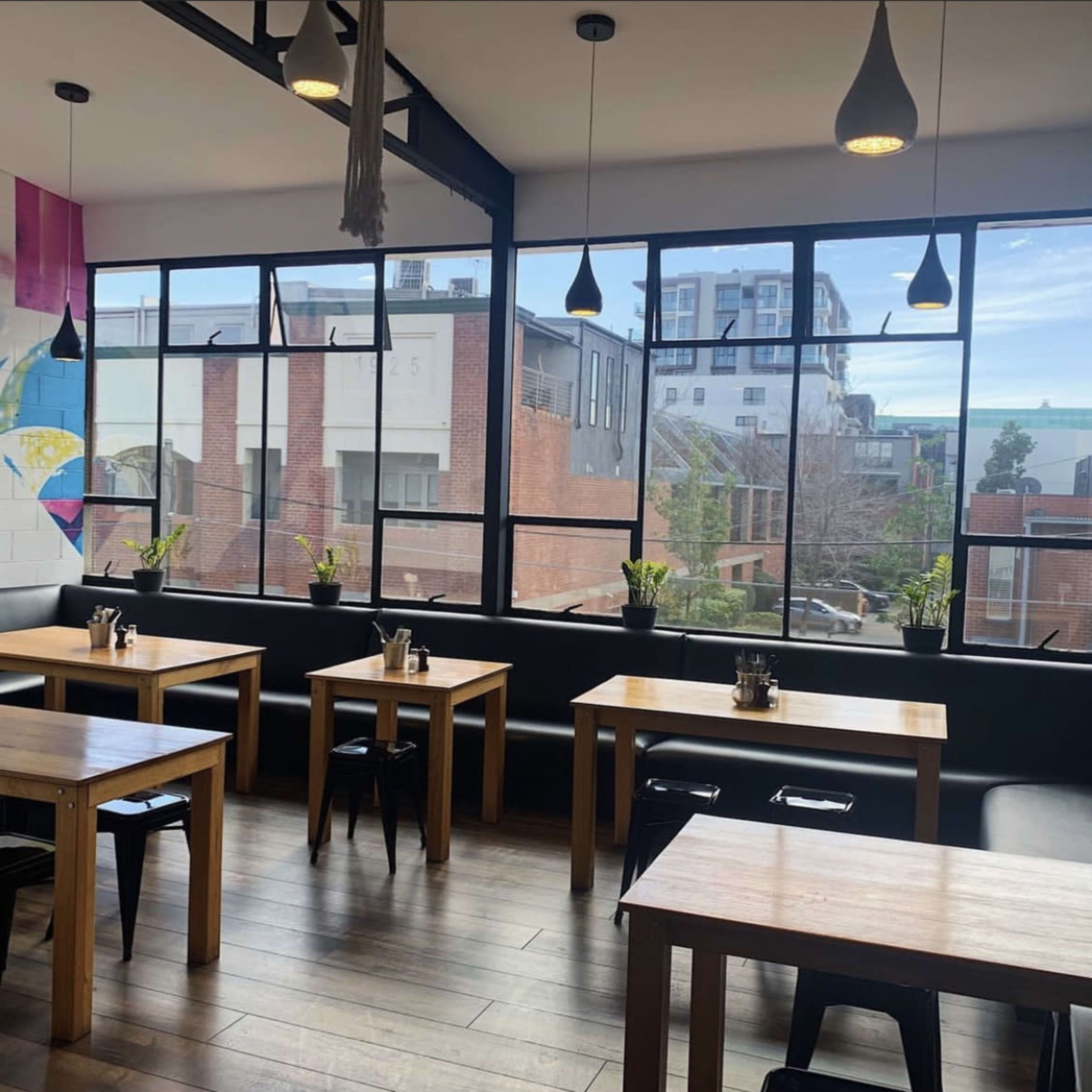 A modern restaurant interior with wooden tables, black chairs, and large windows displaying an urban view.