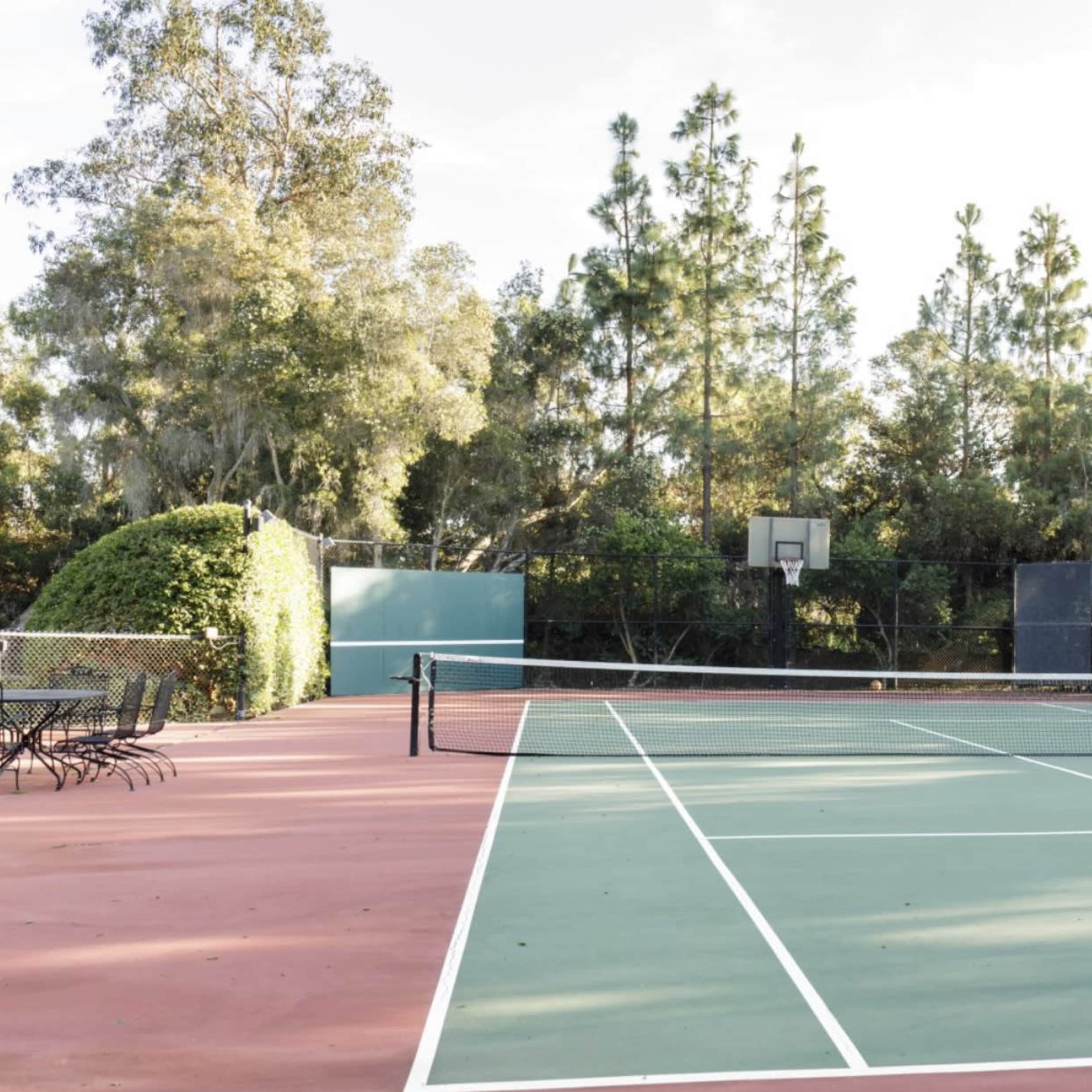 A tennis court with a basketball hoop in the background is surrounded by trees and has a red and green surface.