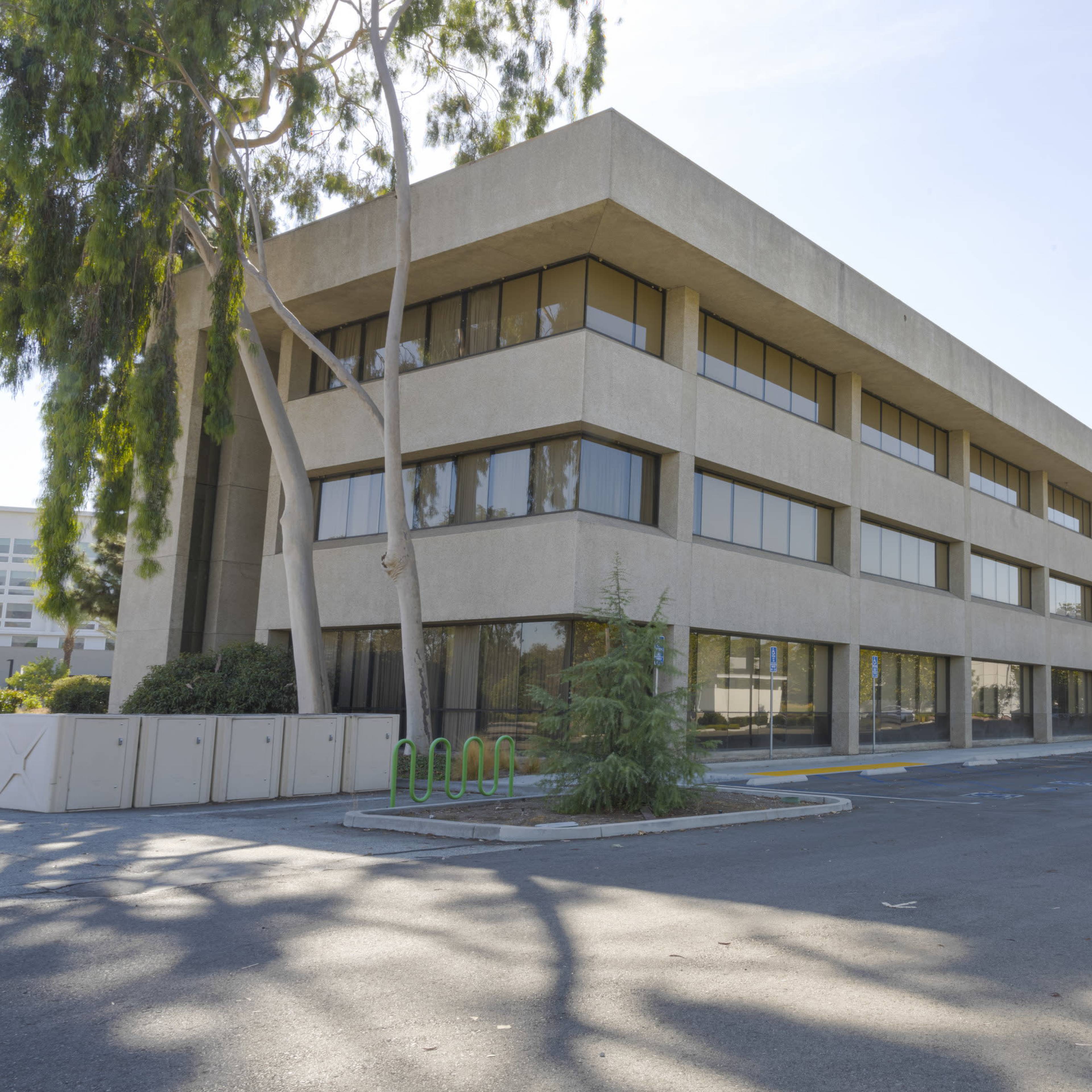 A three-story, modern concrete building with large windows and an adjacent parking area.