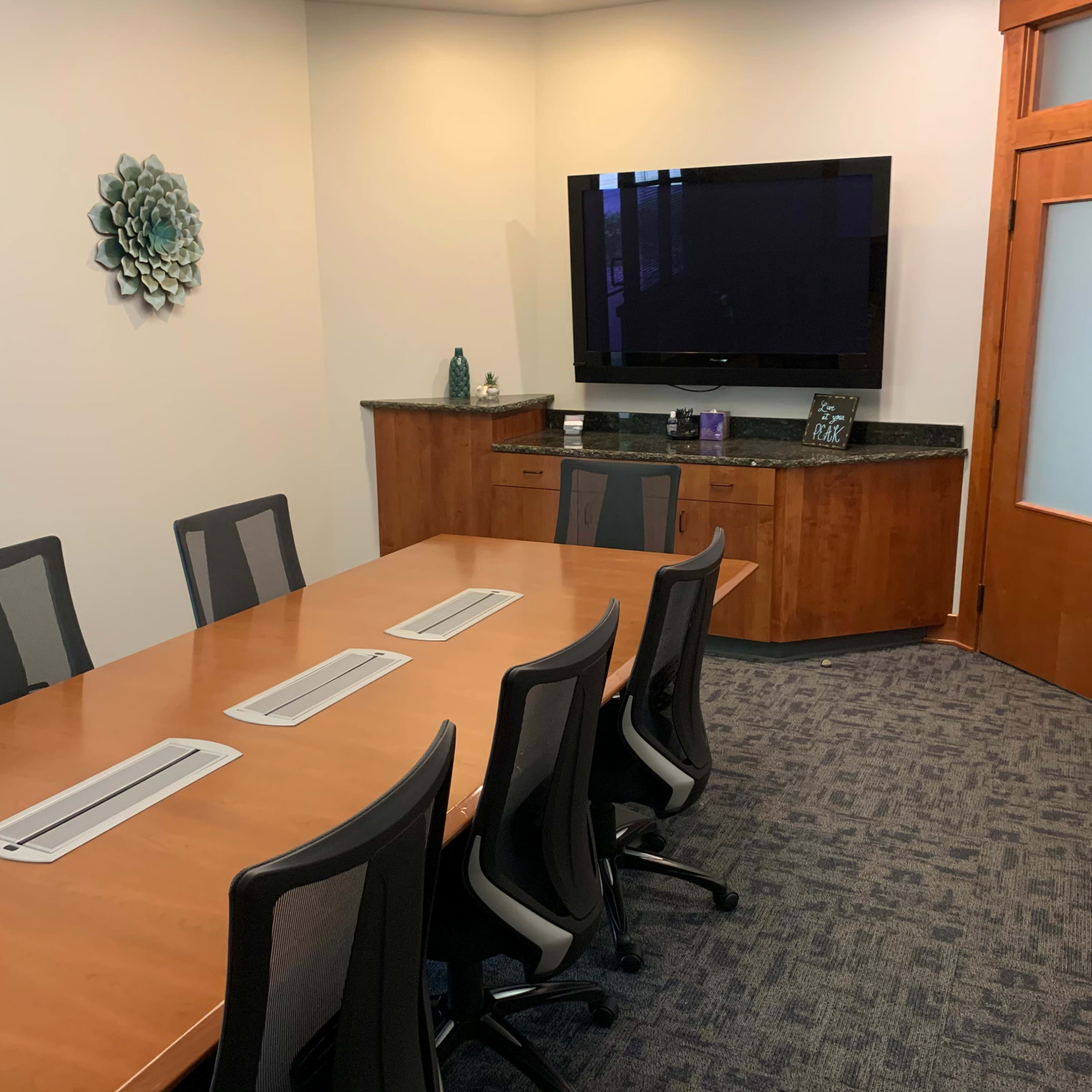 A conference room features a wooden table surrounded by black chairs, a large television mounted on the wall, and a cabinet against the side.