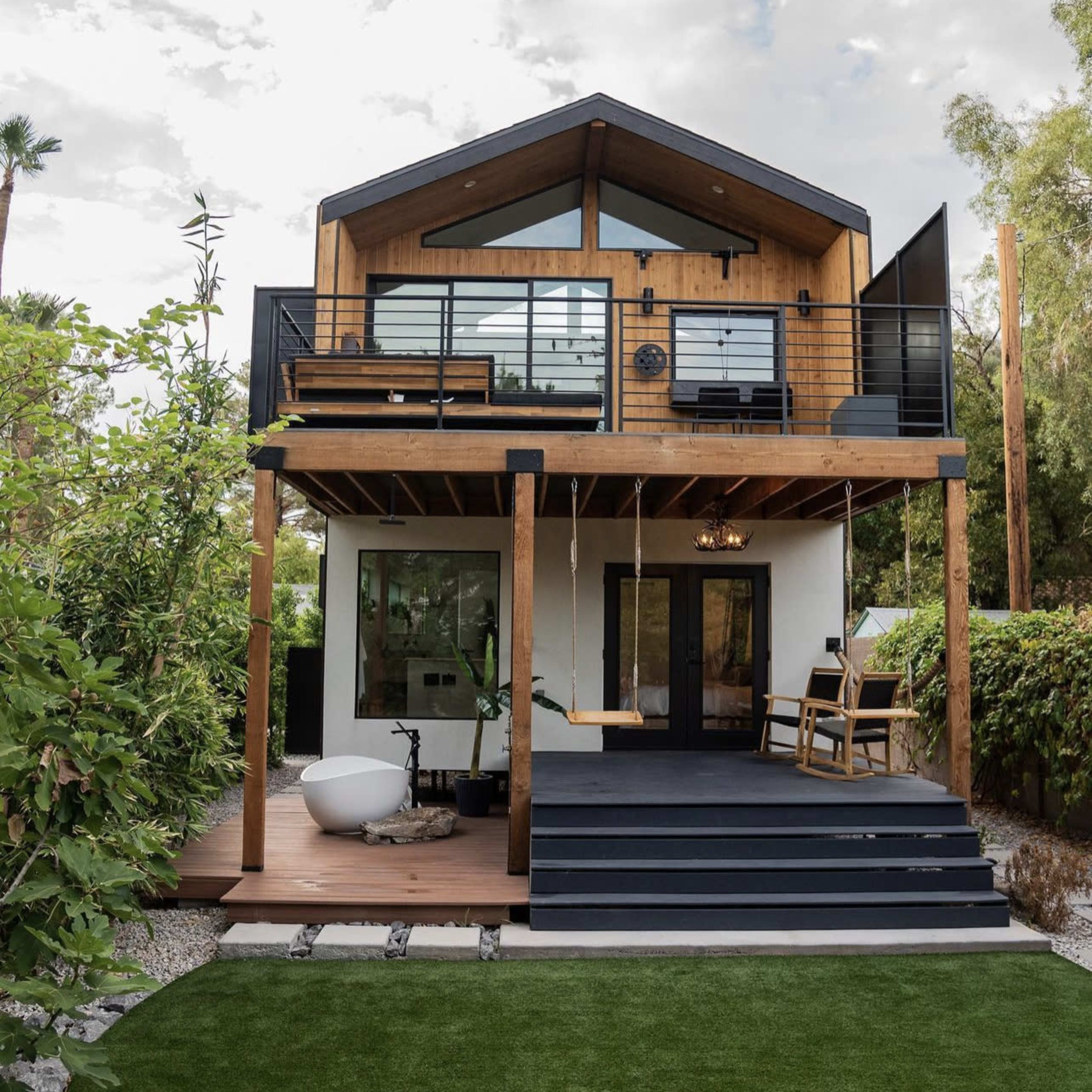 A modern two-story house features a wooden facade, large windows, and a balcony, surrounded by greenery and a landscaped yard.