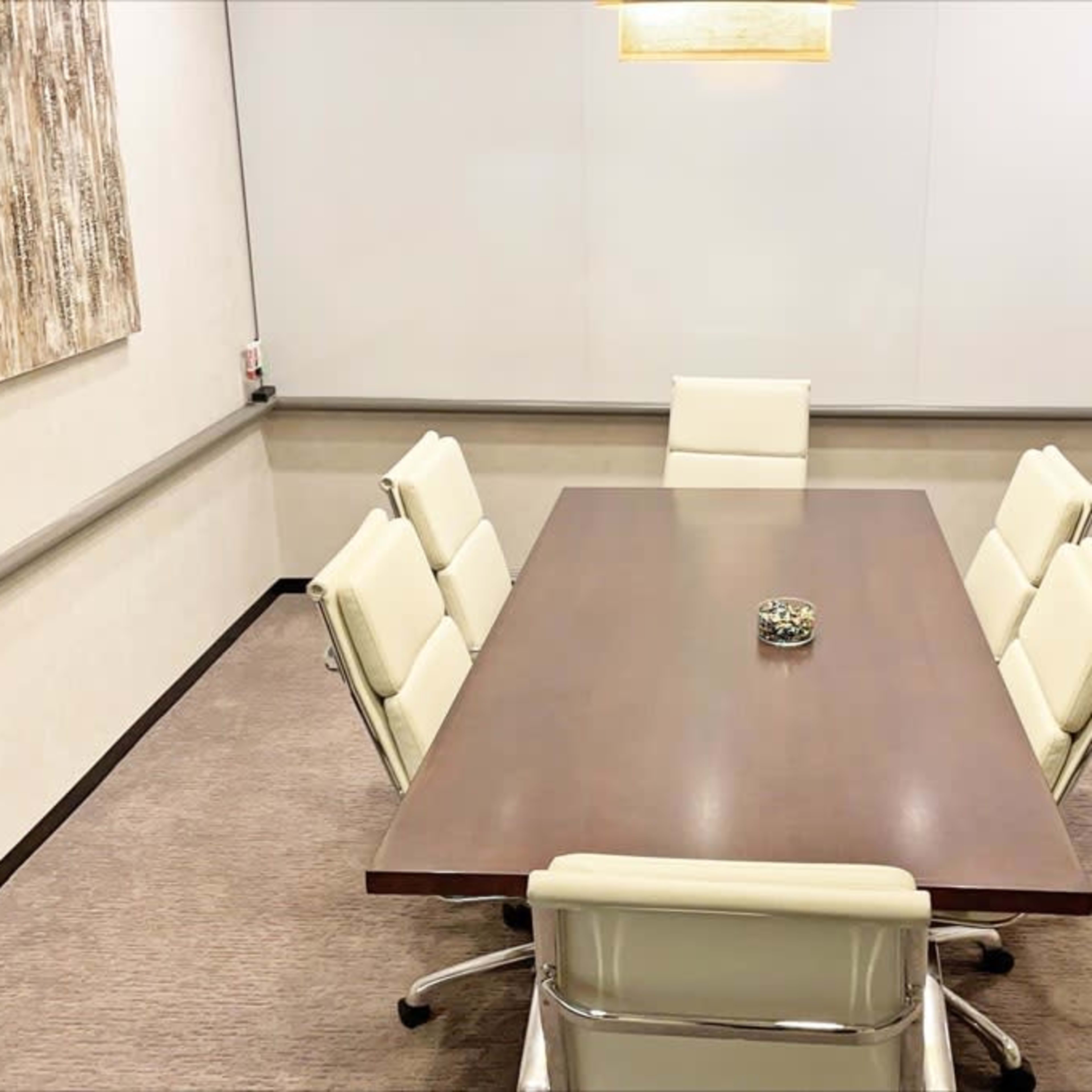 A modern conference room features a large wooden table surrounded by six white chairs, with a decorative item at the center and a television mounted on the wall.