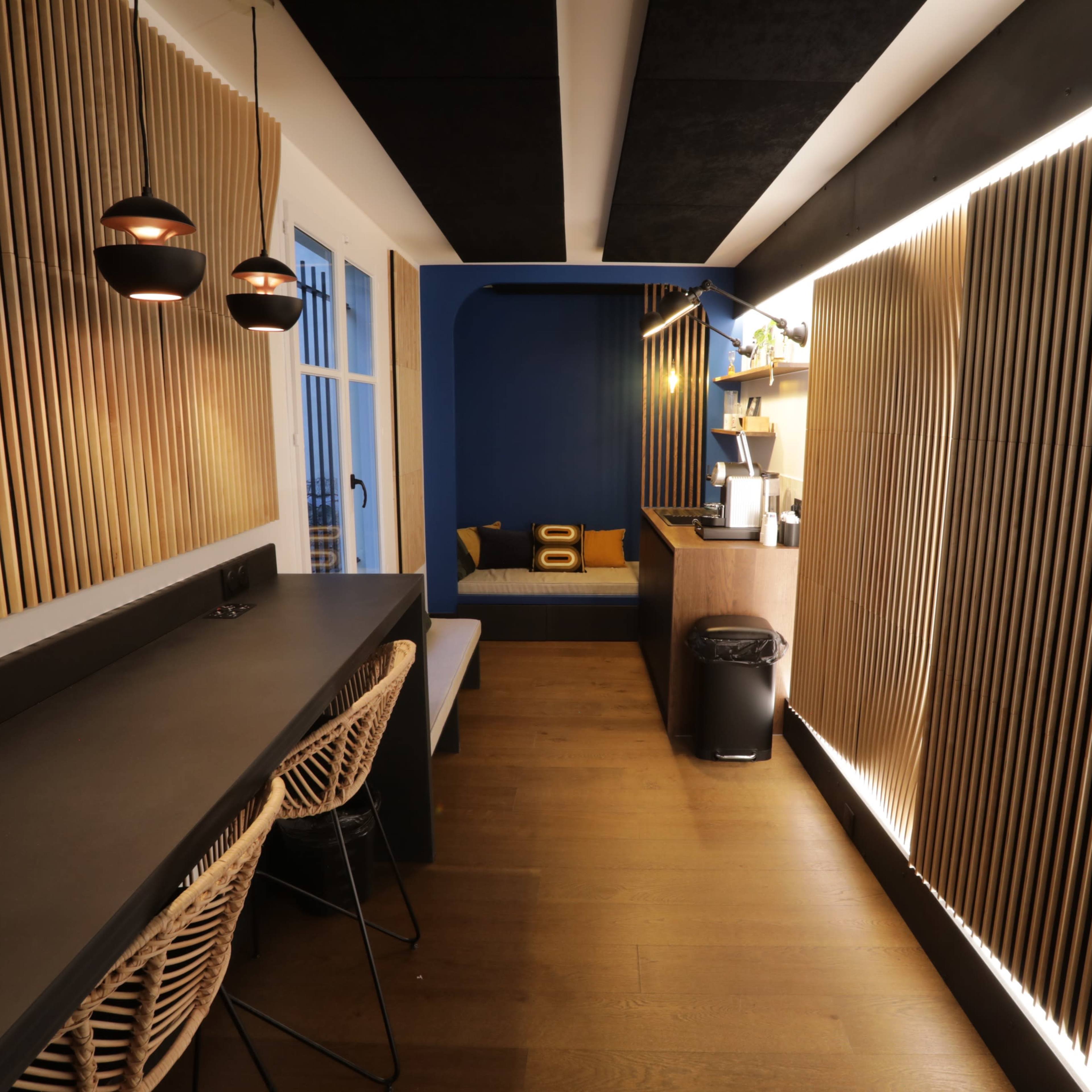 A modern interior space featuring a long black countertop with bar stools, wooden slatted walls, and a cozy seating area illuminated by pendant lights.