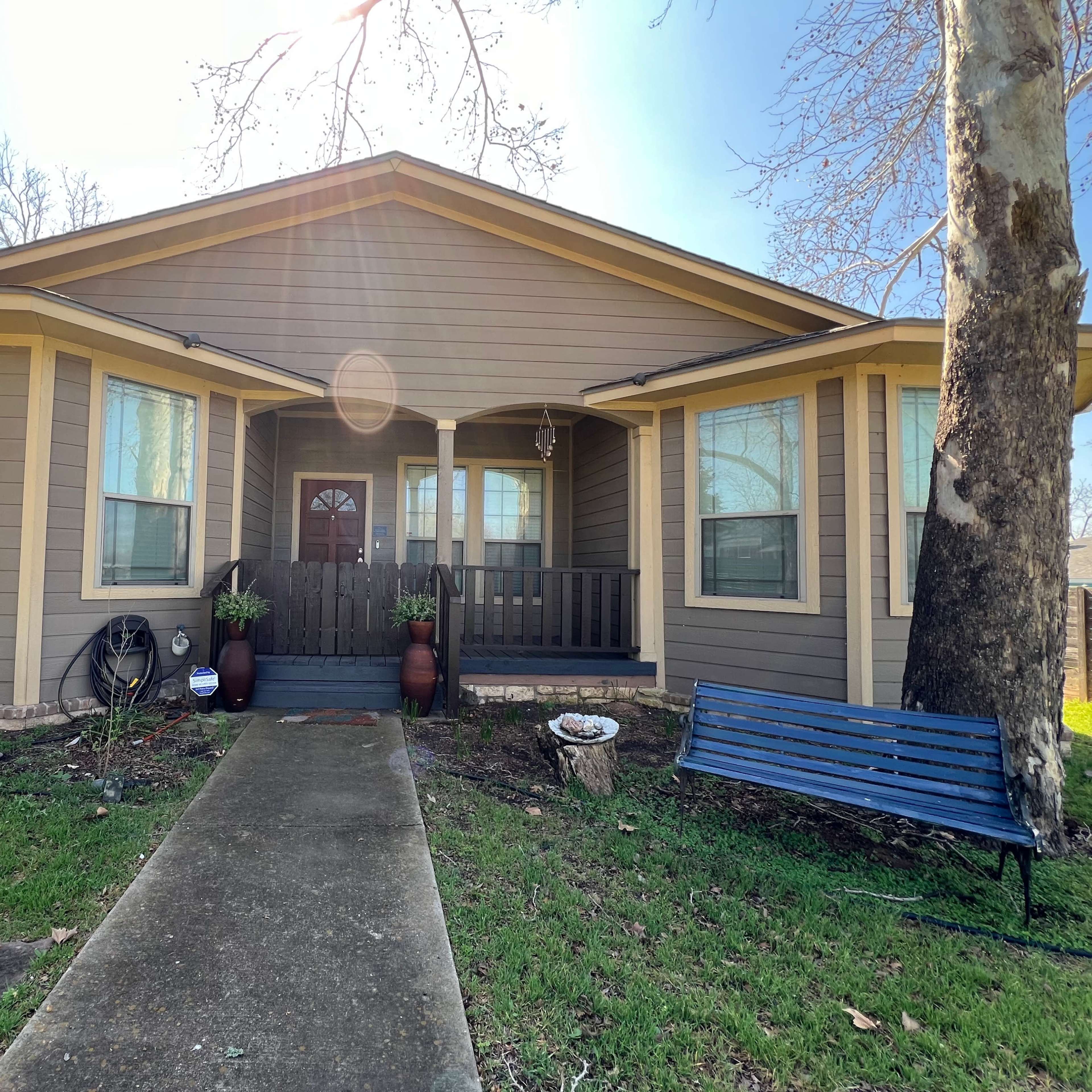 A single-story house with a front porch, large windows, and a pathway leading to the entrance is surrounded by a lawn and a bench.