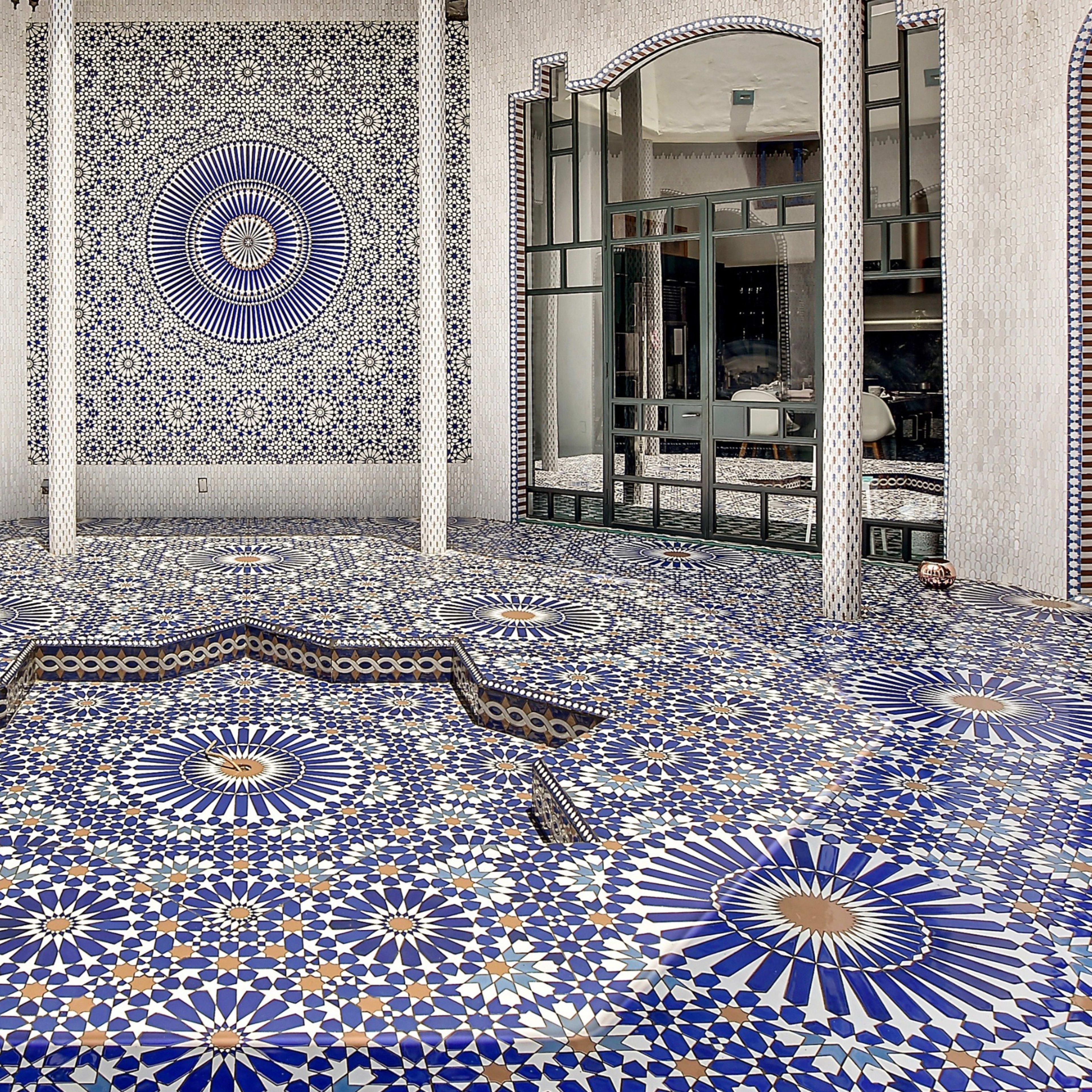 An intricately designed tiled floor and wall featuring geometric patterns in blue, white, and yellow in a spacious, brightly lit room with large windows.