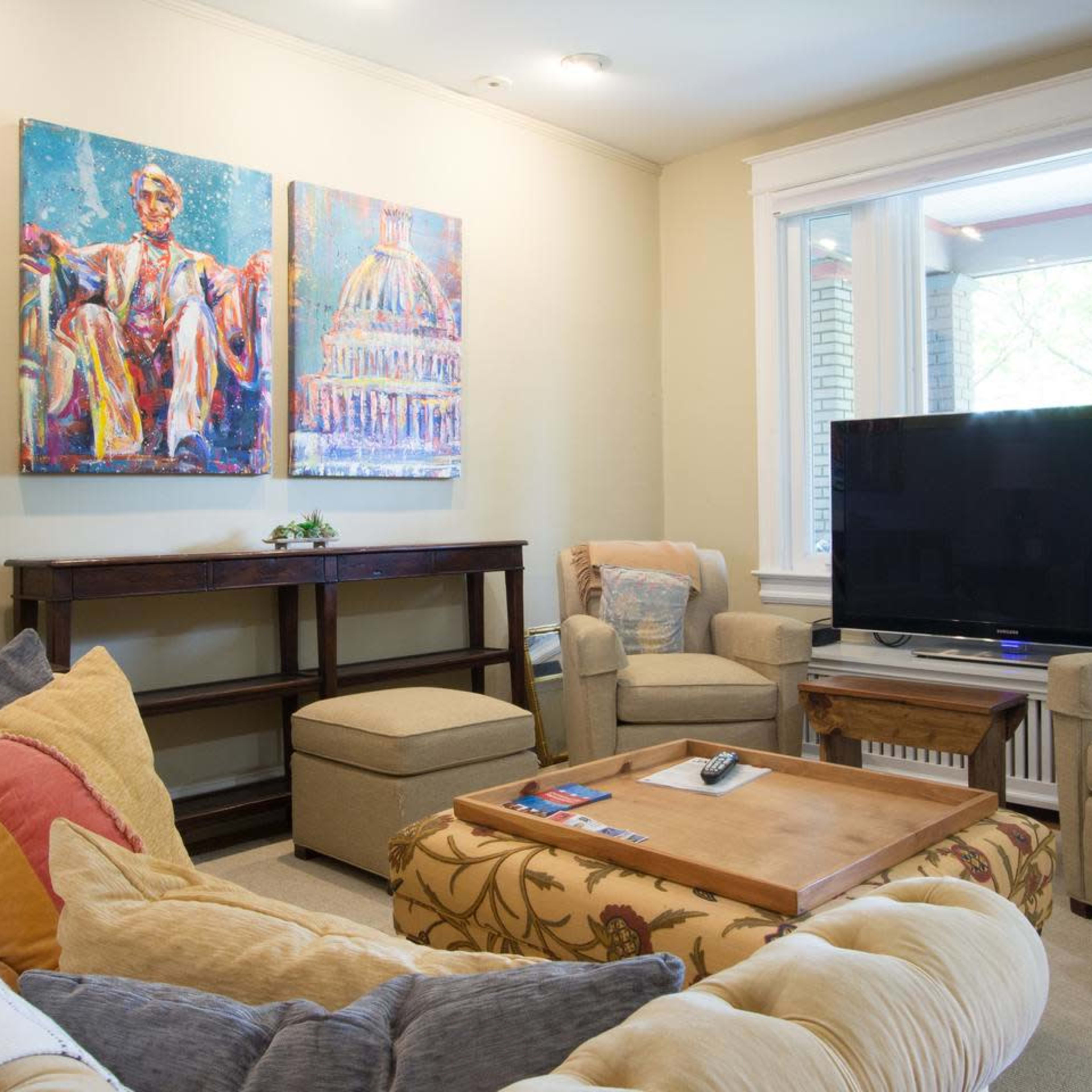 A cozy living room featuring two colorful paintings, a large television, a wooden coffee table, and comfortable seating arrangements.