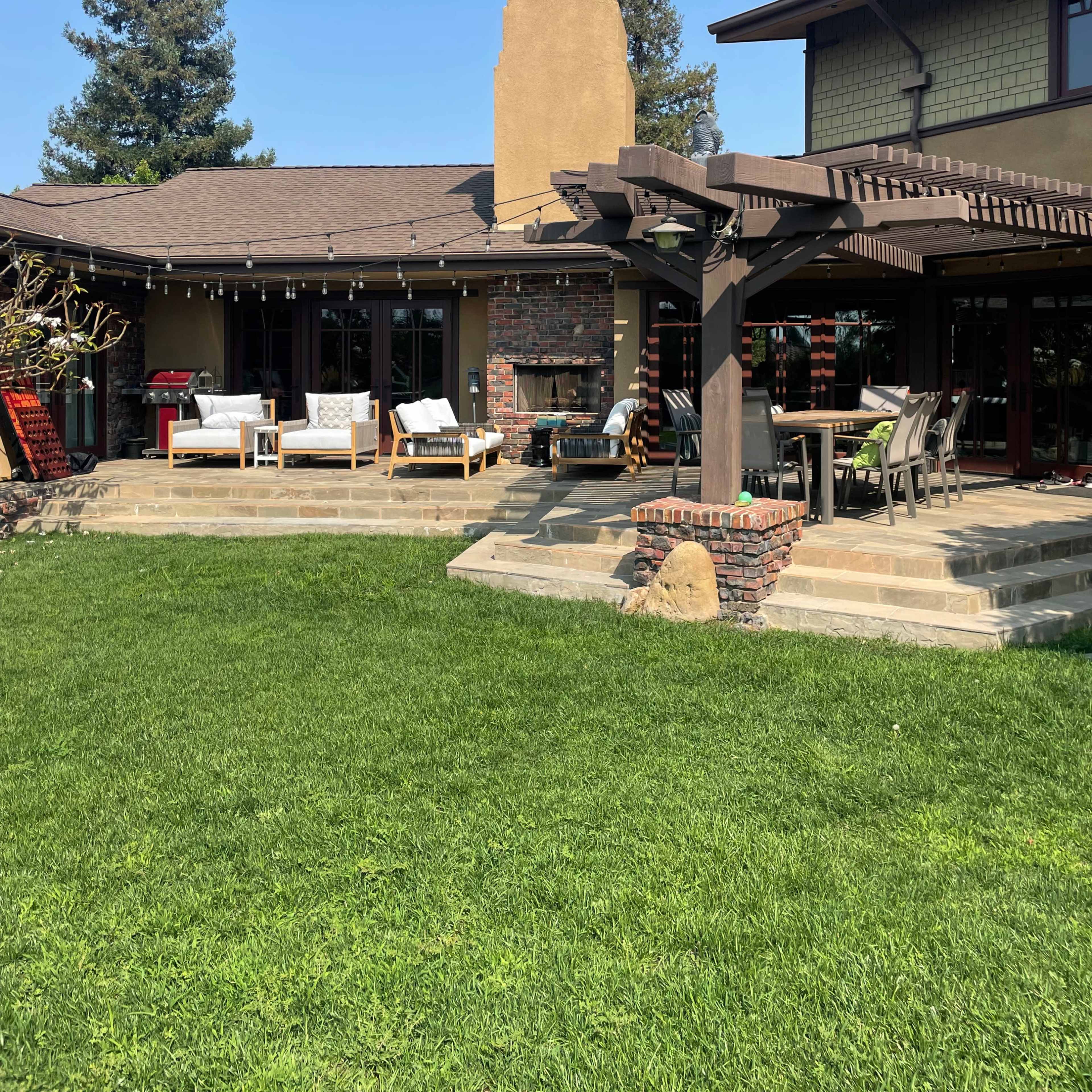 A modern house with a wooden pergola and outdoor seating is set against a backdrop of green lawn and clear blue sky.