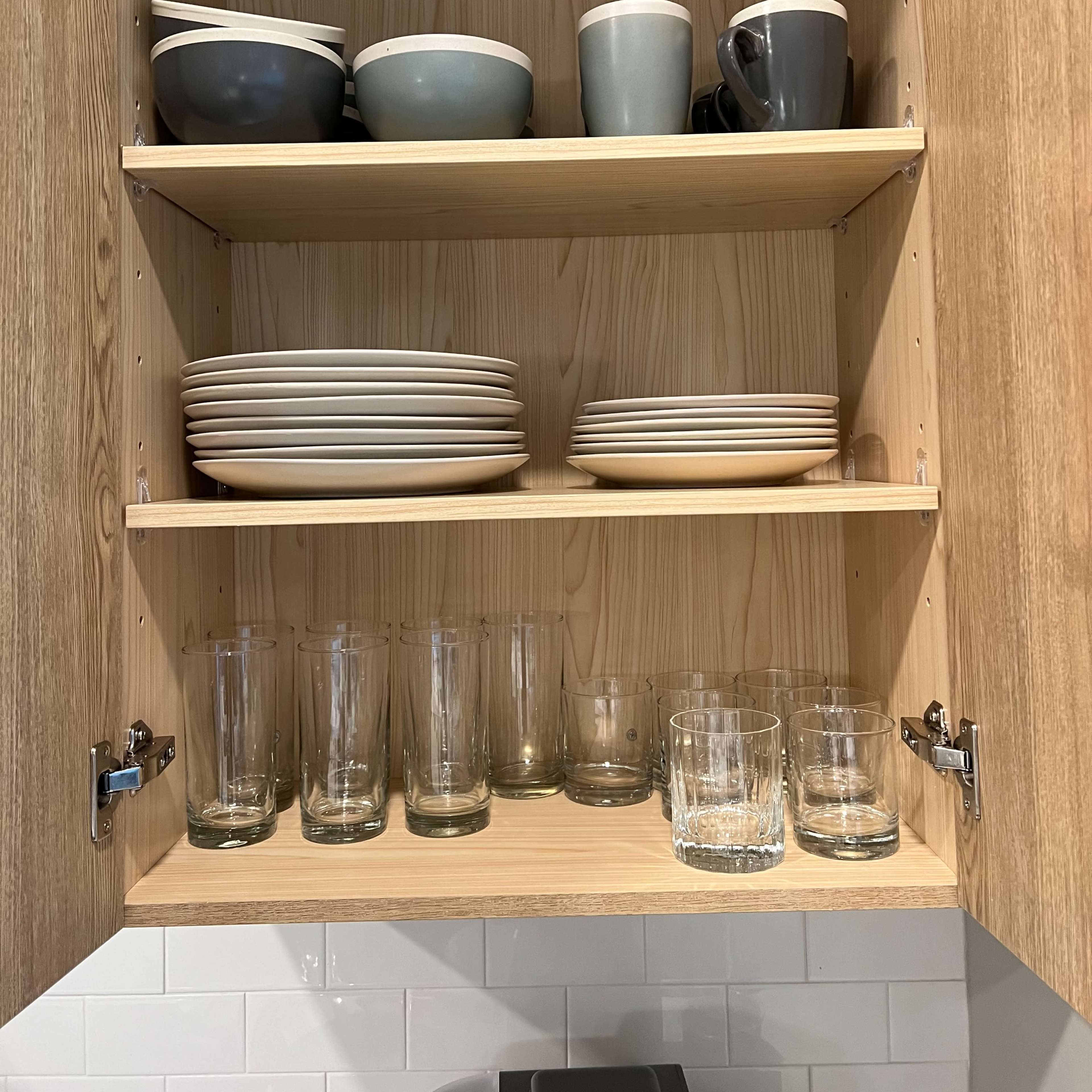 A wooden kitchen cupboard with three shelves containing a variety of bowls, mugs, plates, and drinking glasses.