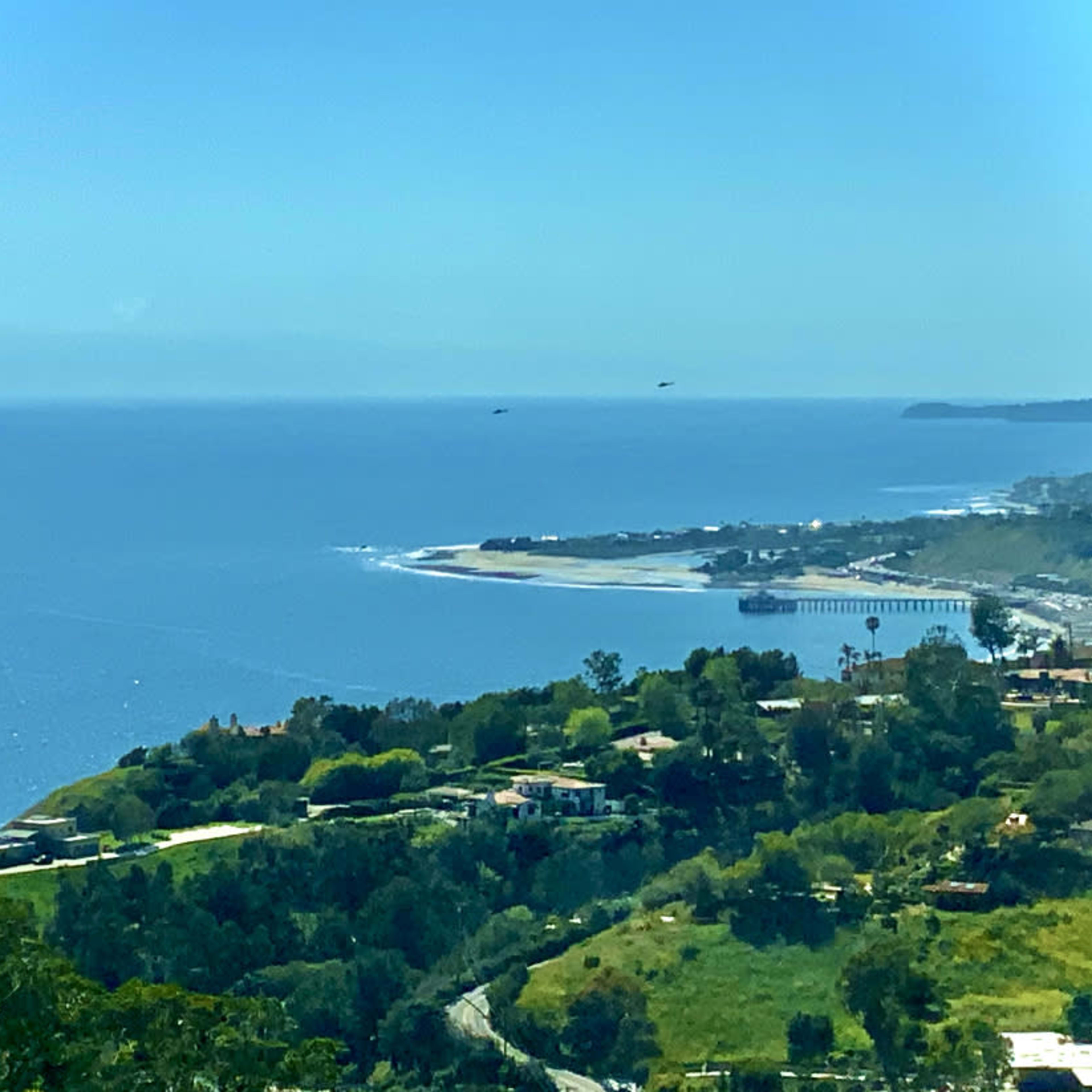 A coastal view featuring a sandy beach, a pier, and lush greenery in the foreground.
