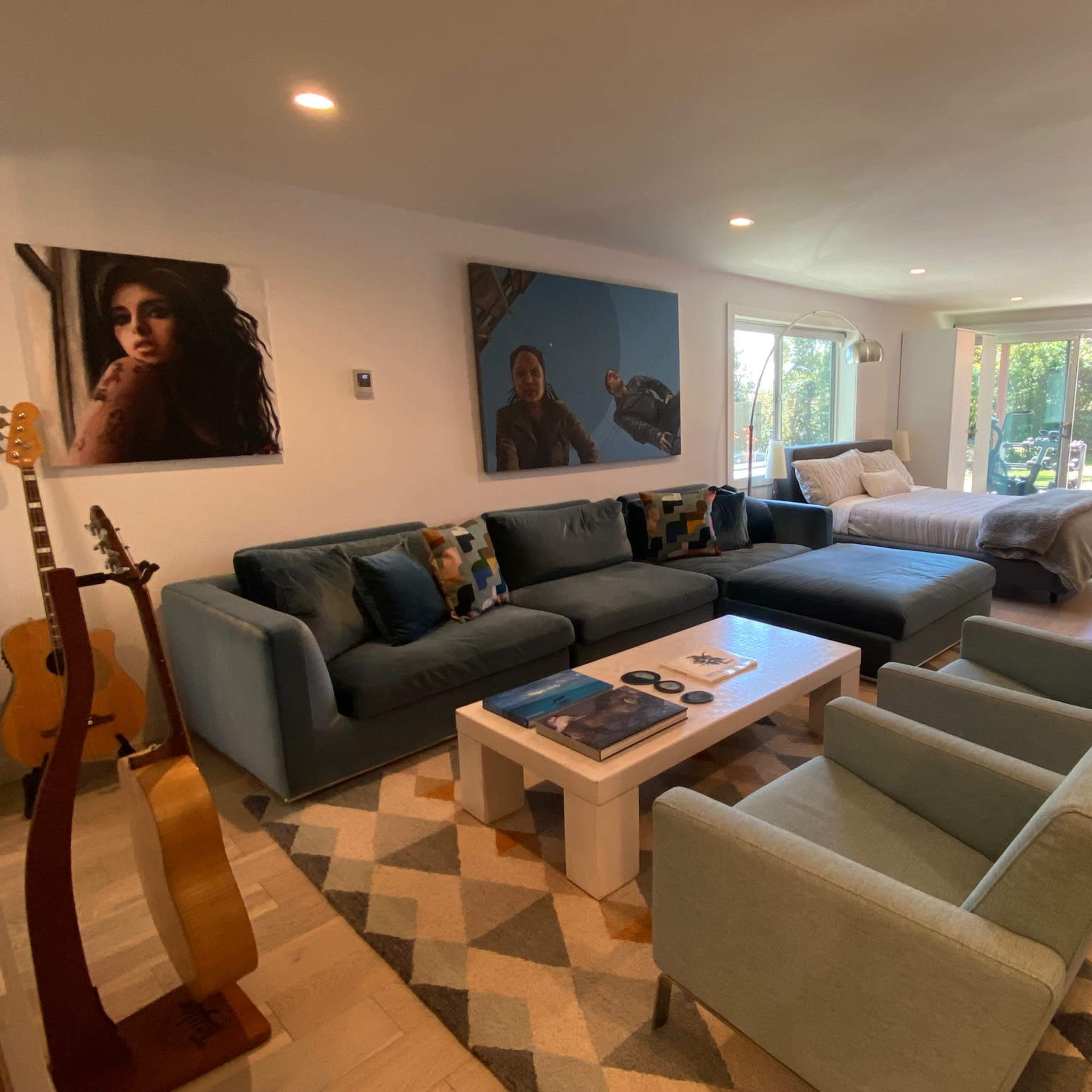 A spacious living room features a sectional sofa, decorative pillows, a coffee table, artwork on the walls, and large windows that open to a garden view.