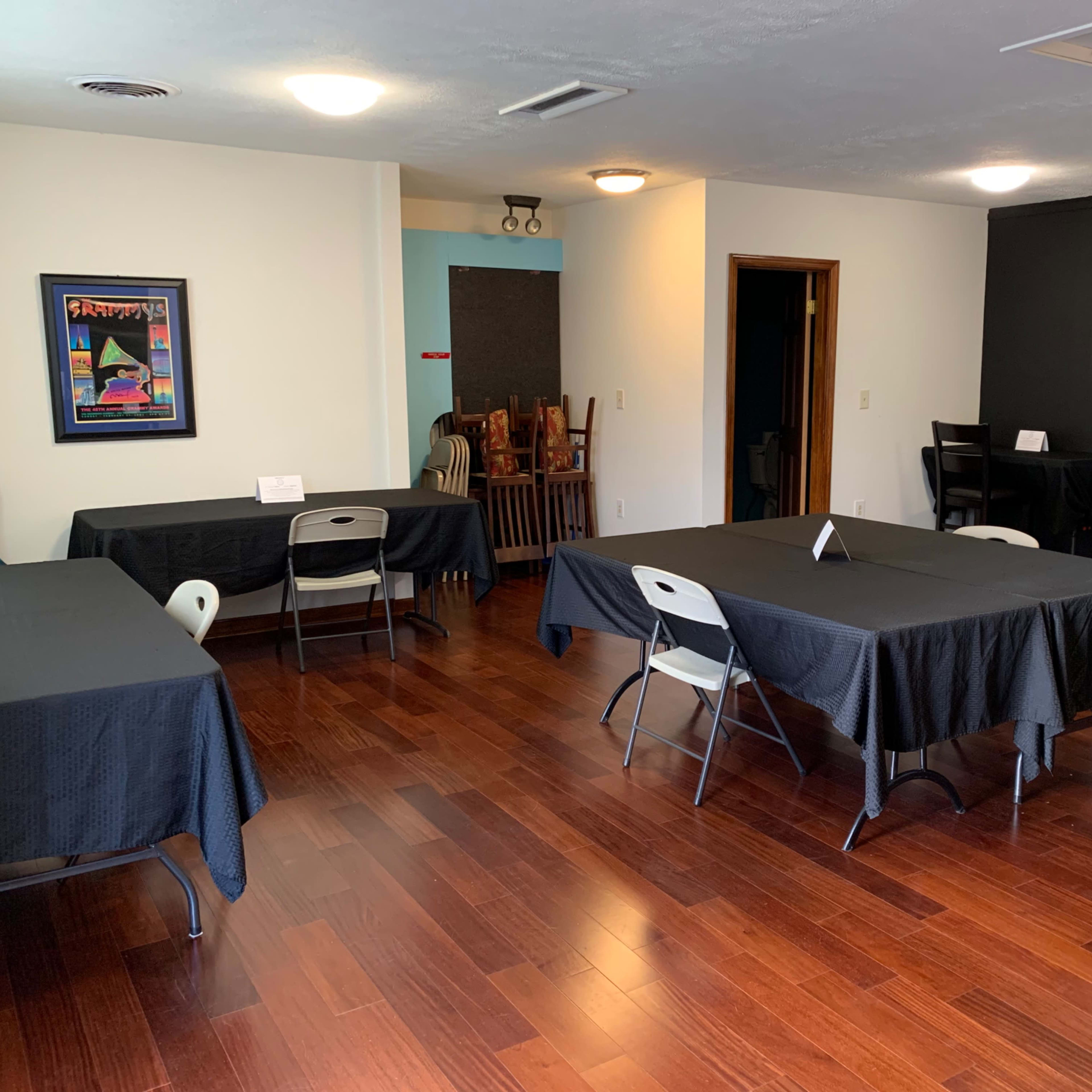 The room features several tables covered with black cloth, arranged with chairs around them, and a blank wall where a projector screen could be placed.