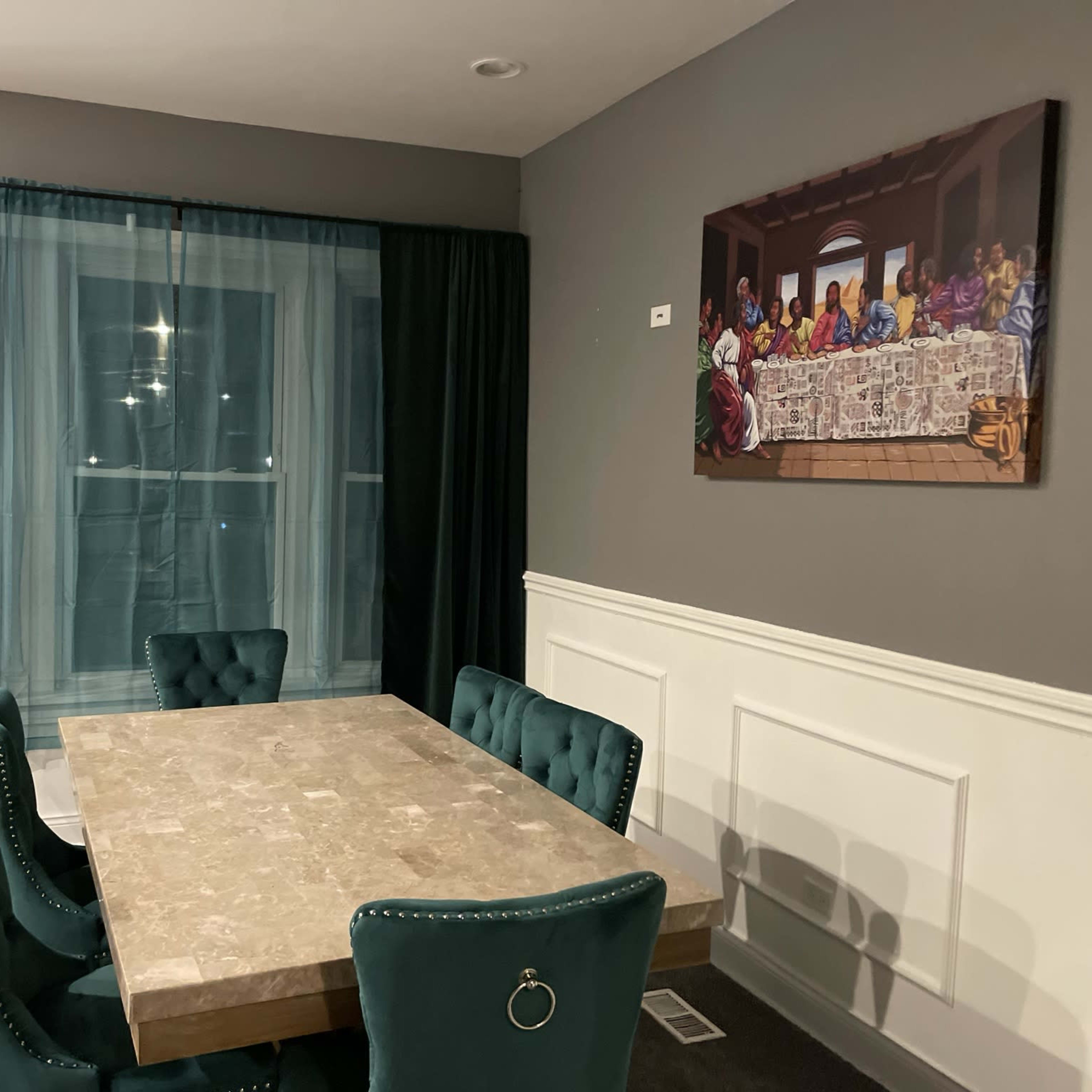 A dining room features a stone table surrounded by upholstered chairs and a large window with dark curtains, while a painting hangs on the wall depicting a group gathered around a long table.