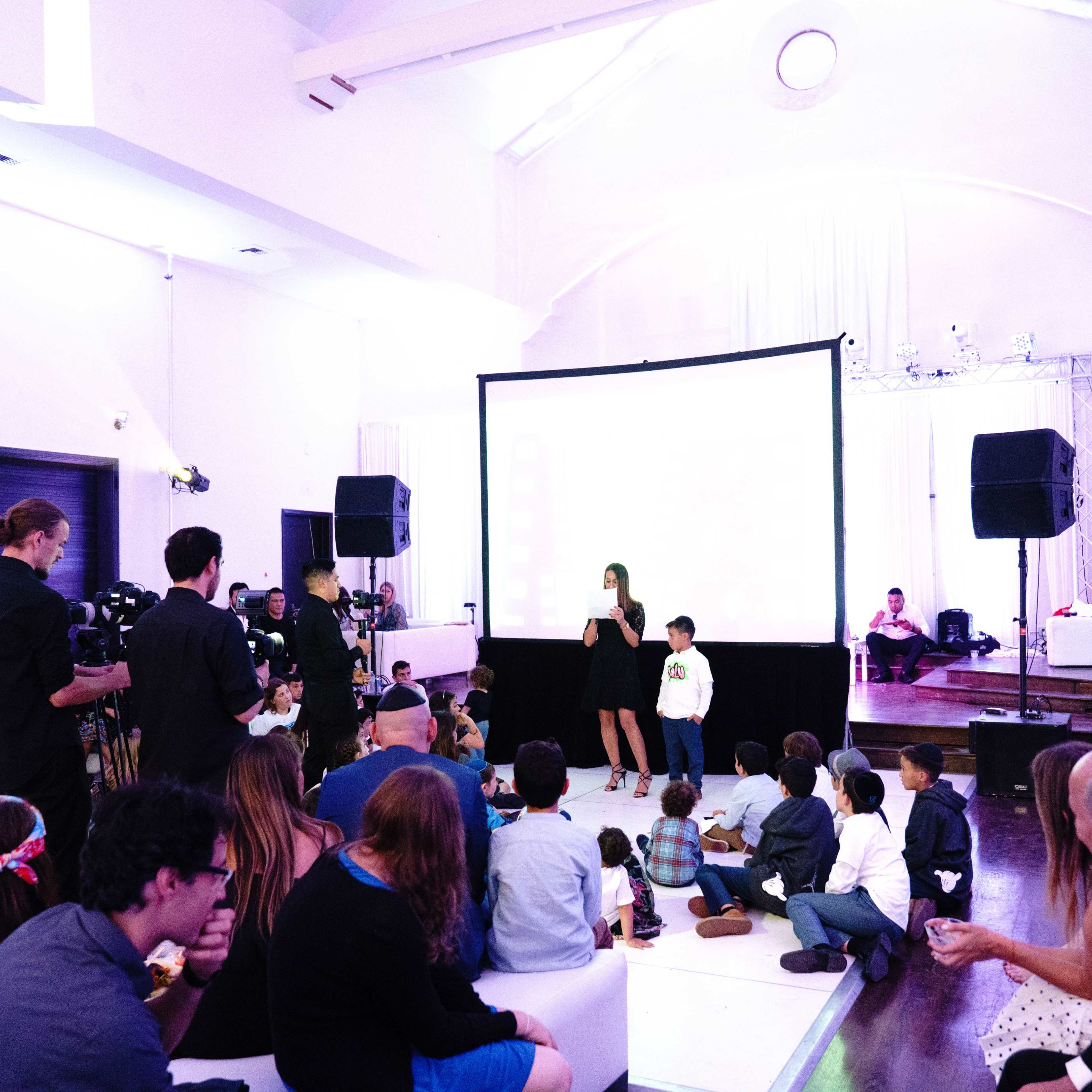 A woman and a child are speaking on stage in front of an audience seated on the floor in a brightly lit event space.