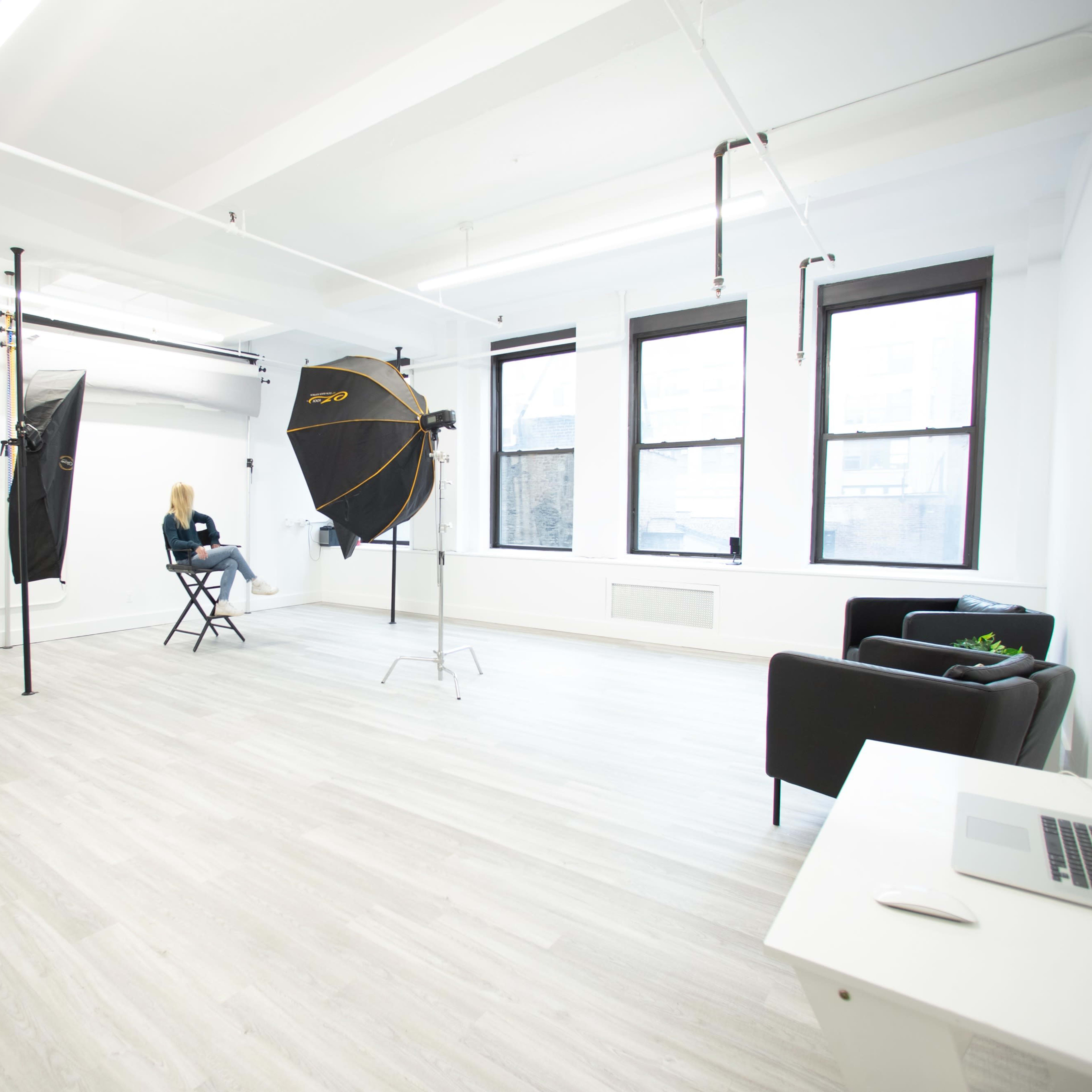 A bright photography studio with two large softboxes, a chair, and a small black sofa, all arranged in a minimally decorated space.