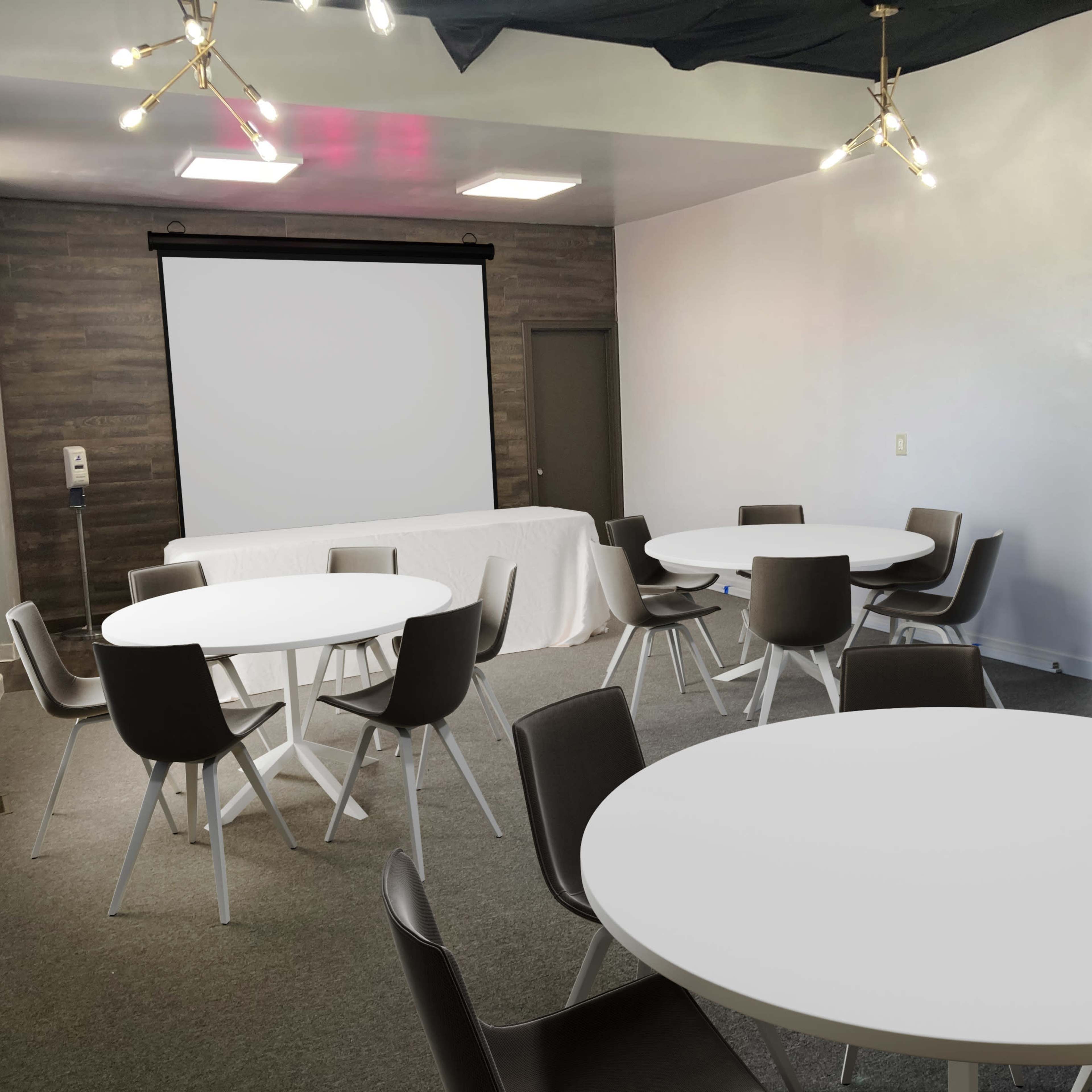 A modern meeting room with round tables and chairs, a projector screen on the wall, and a decorative mirror.