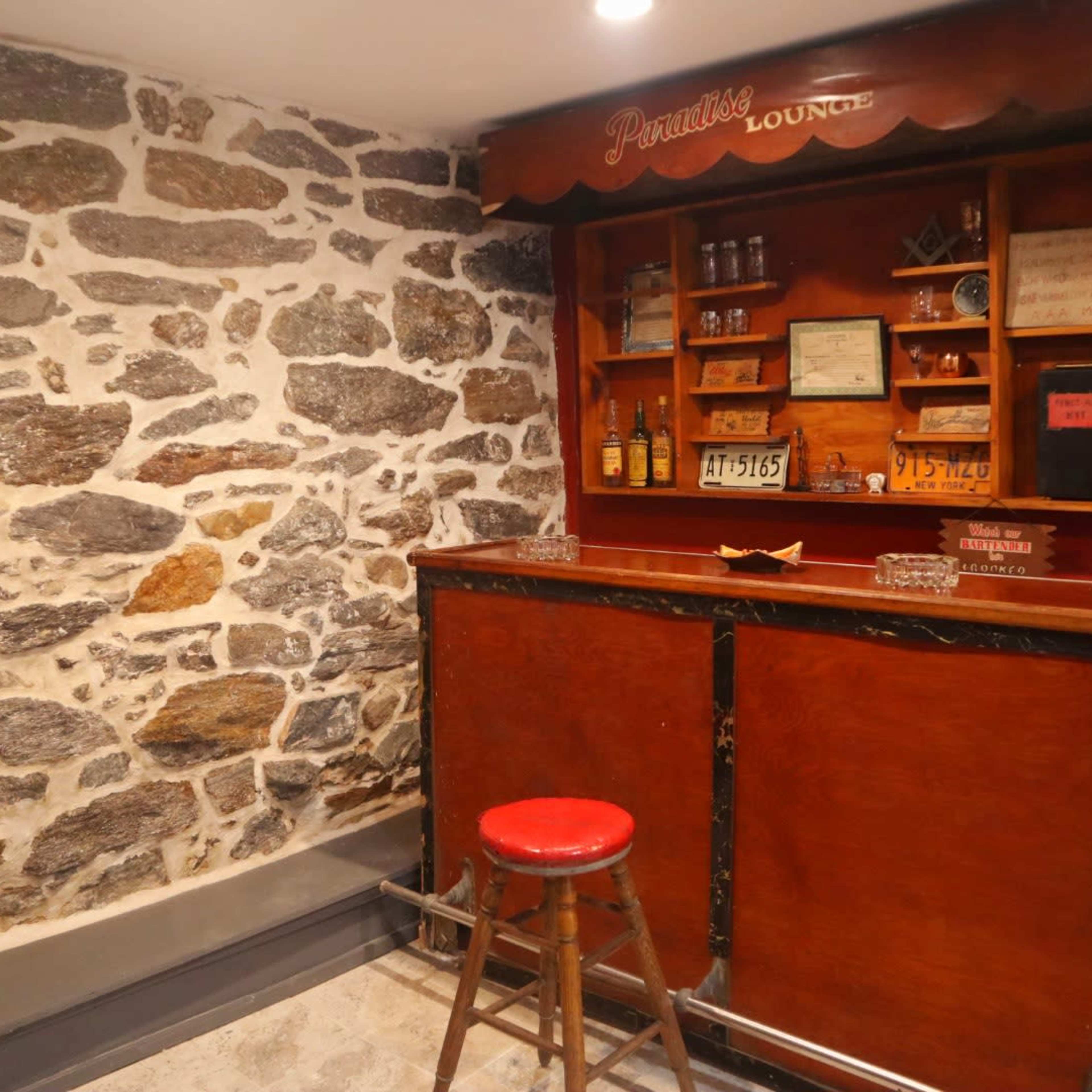 A small bar area with a wooden counter and a red stool, situated against a backdrop of a rustic stone wall.