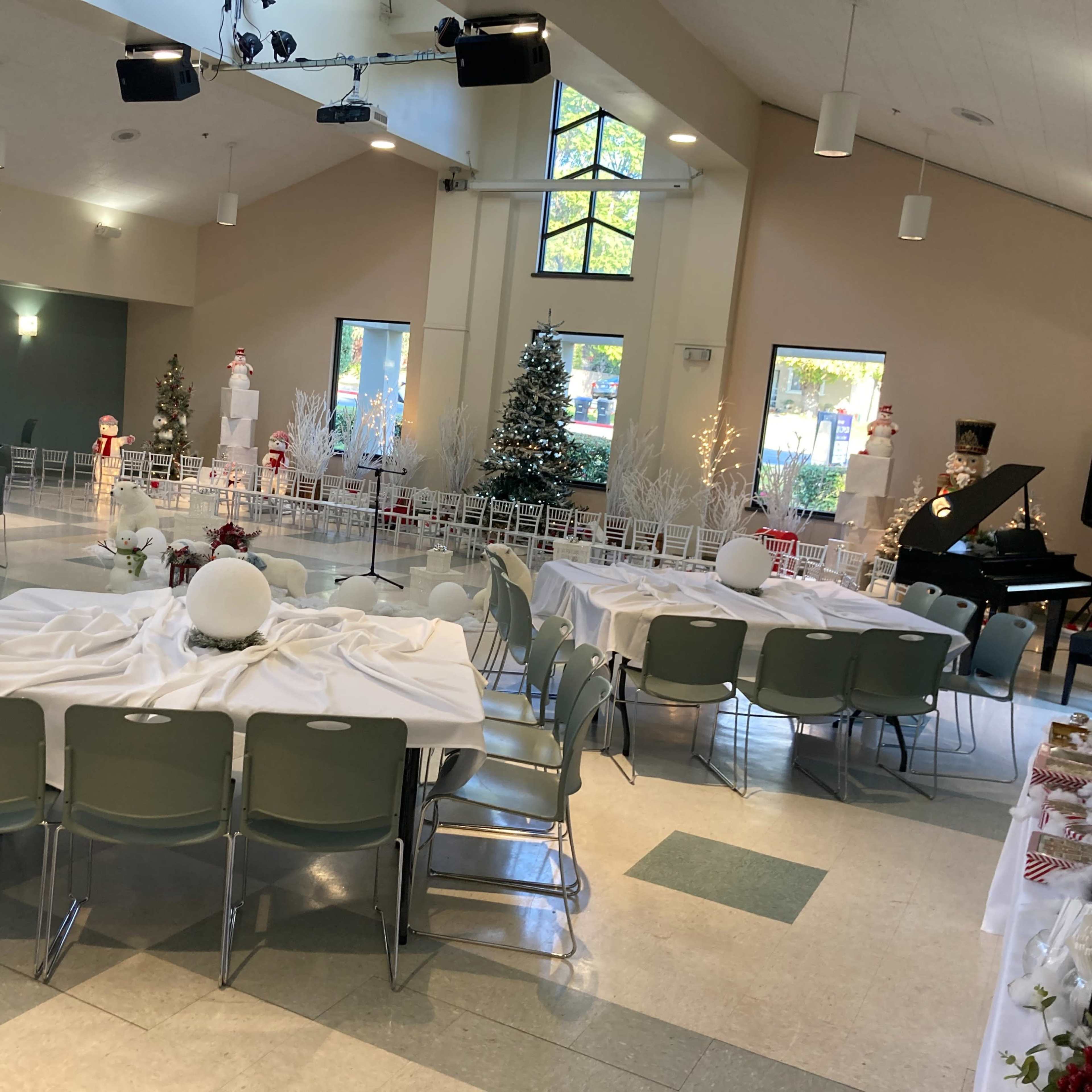 A large indoor event space set up for a gathering, featuring round tables covered with white tablecloths, decorated with holiday-themed centerpieces, and a piano positioned against a wall.