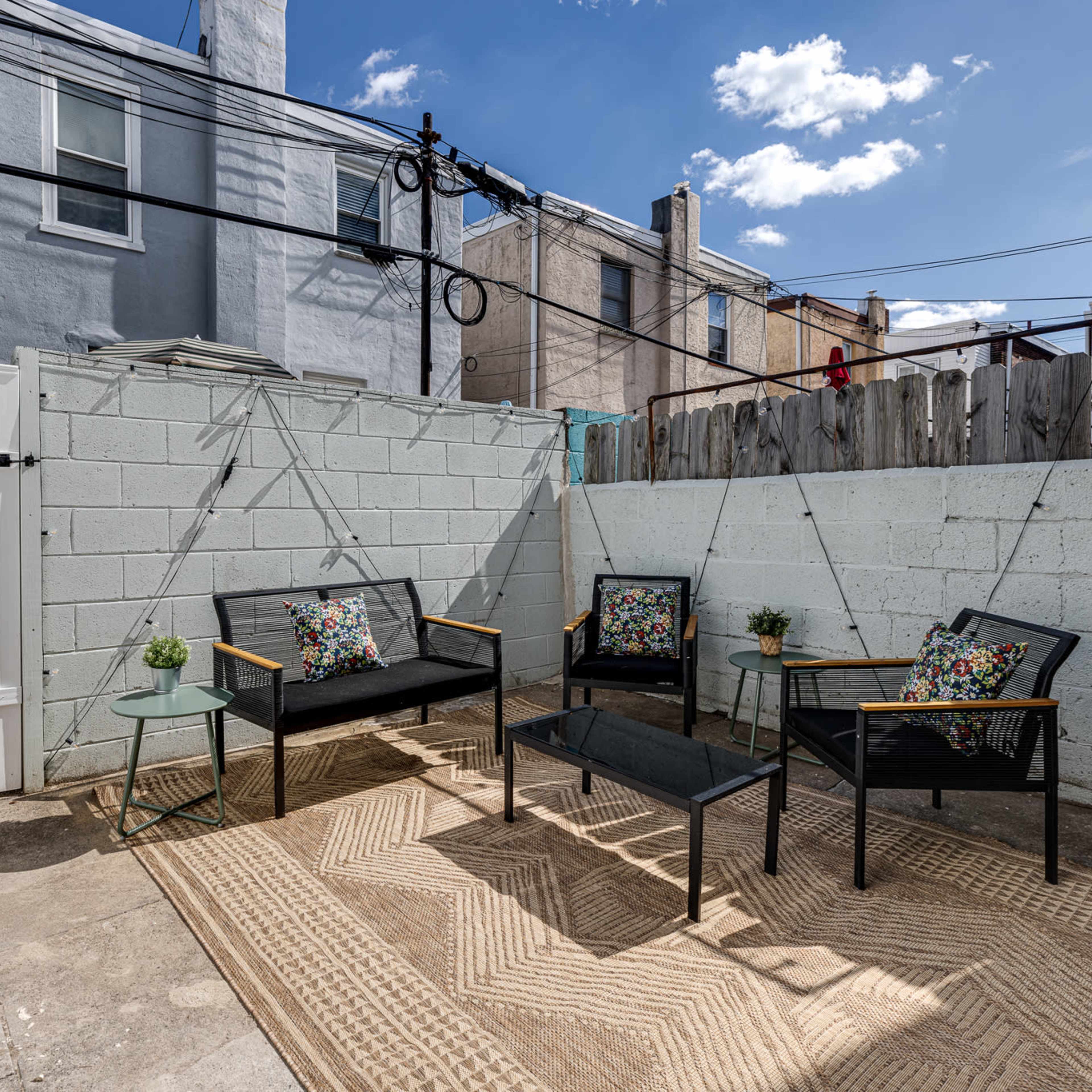 A small outdoor patio area with black seating, a glass table, a grill, and a textured rug, surrounded by a concrete wall and residential buildings in the background.