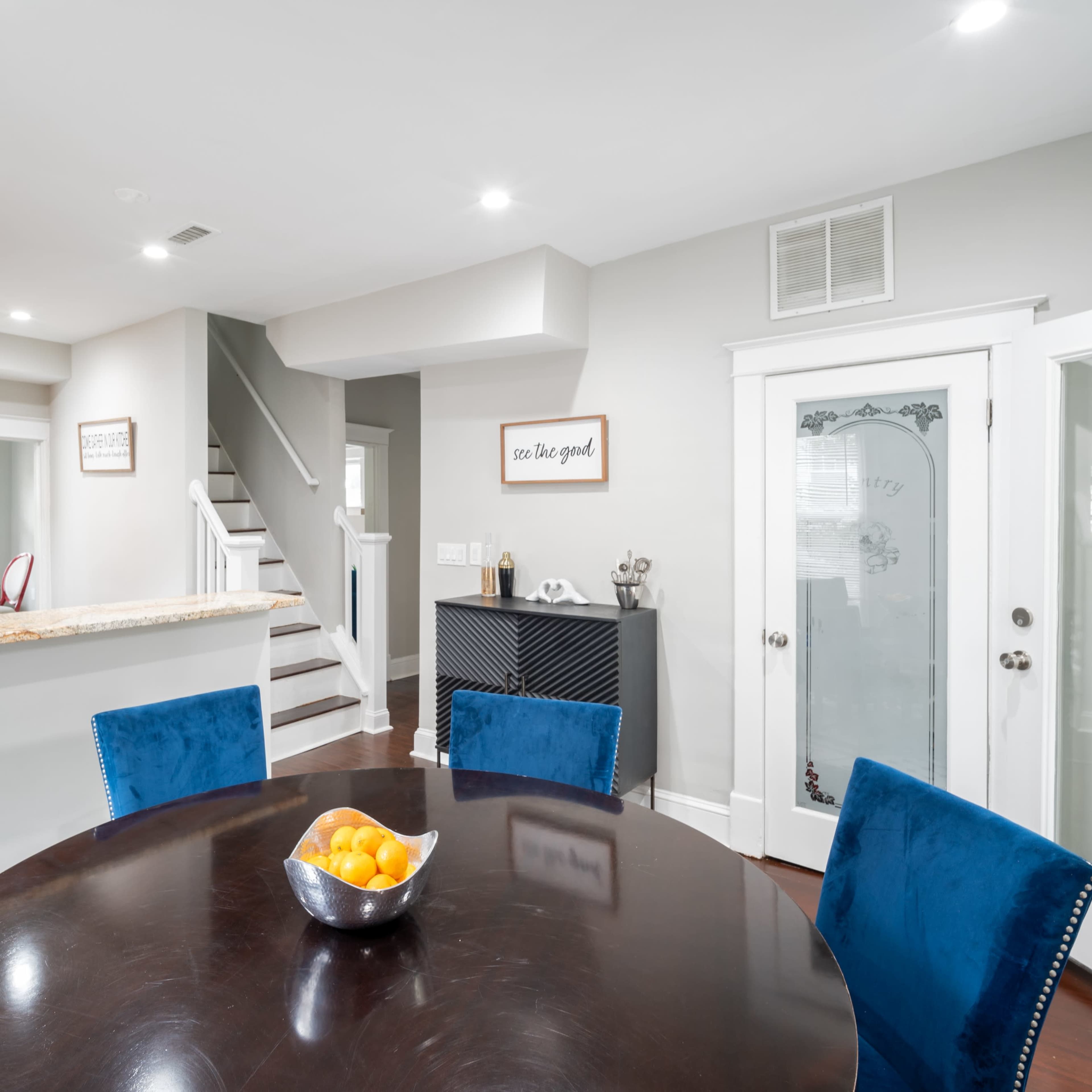A modern dining area with a round table, blue upholstered chairs, and a bowl of oranges, adjacent to a kitchen and a staircase.