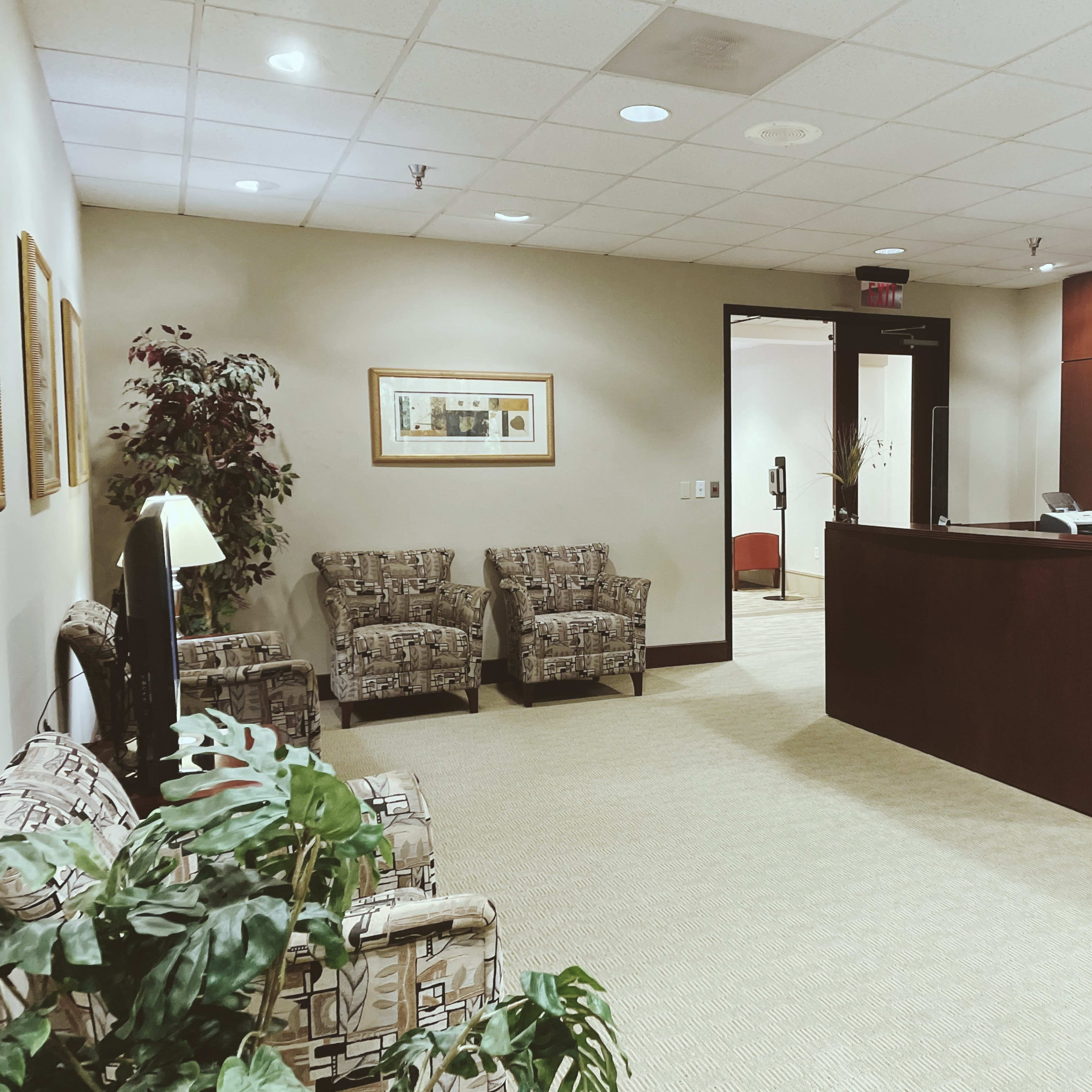 A reception area with two patterned chairs, a plant, and a wooden desk at the front of a well-lit office space.
