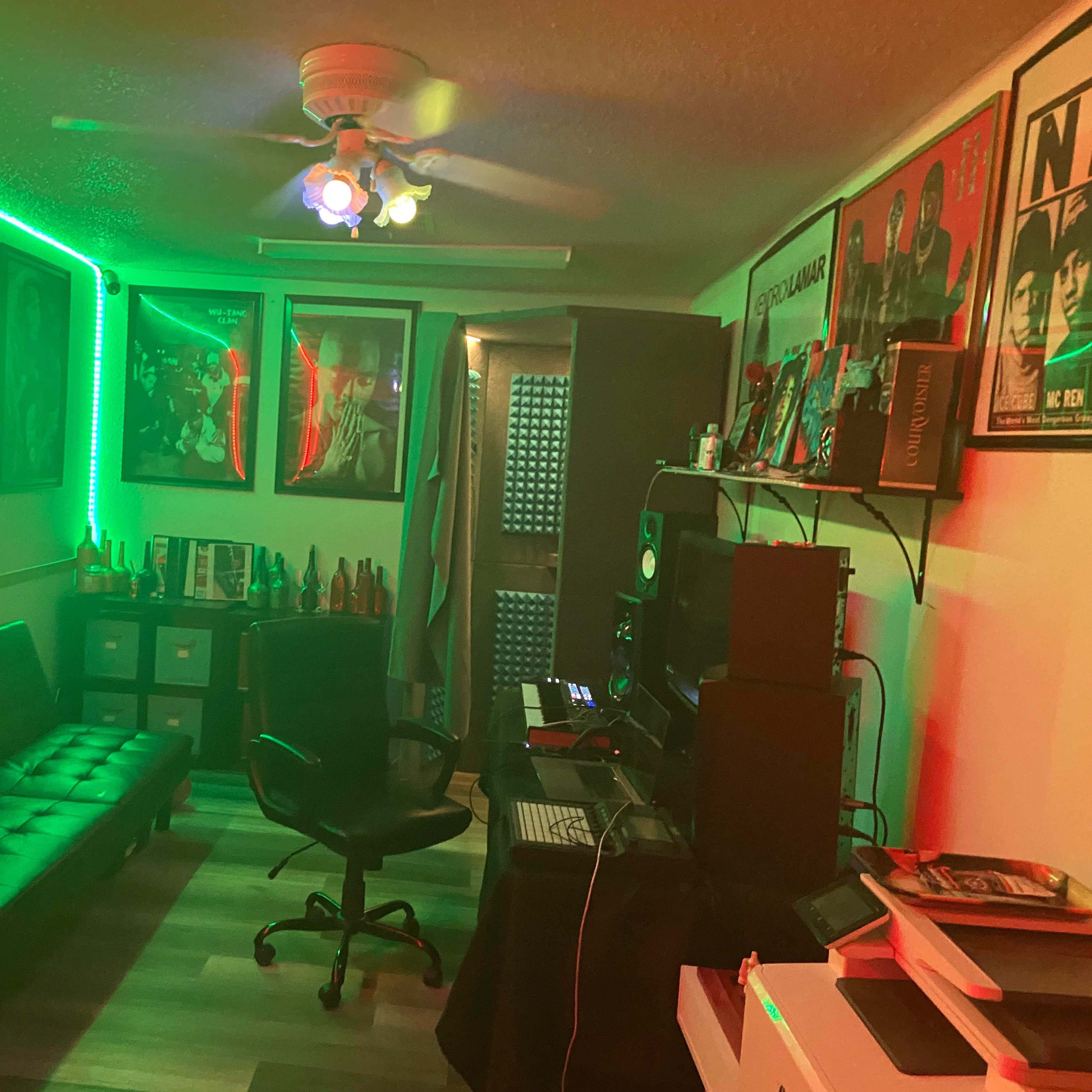 A dimly lit room with green LED lights, featuring a black sofa, a desk with audio equipment, and framed posters on the walls.