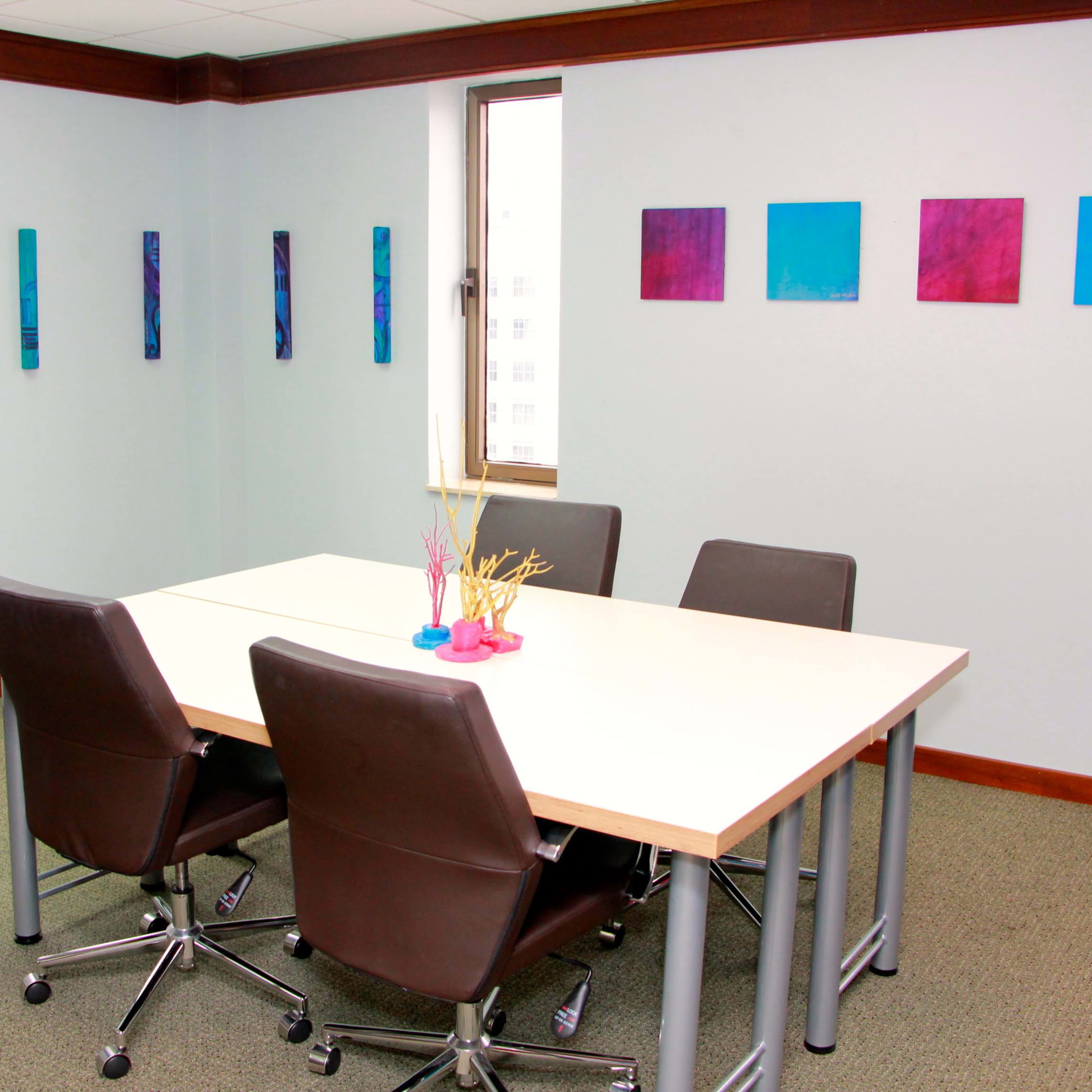 A small conference room features a rectangular table surrounded by four chairs, with colorful paintings on the walls.
