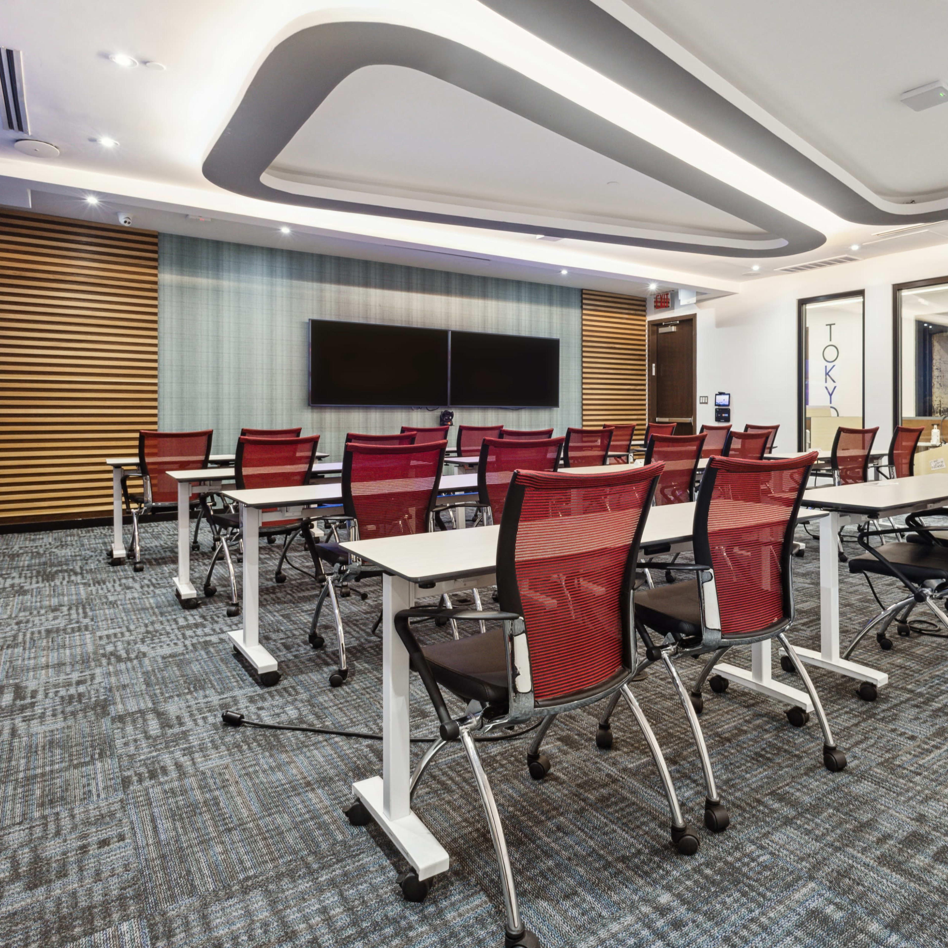 A modern conference room with rows of red and white chairs arranged around tables, a large screen on one wall, and decorative features in a corporate setting.