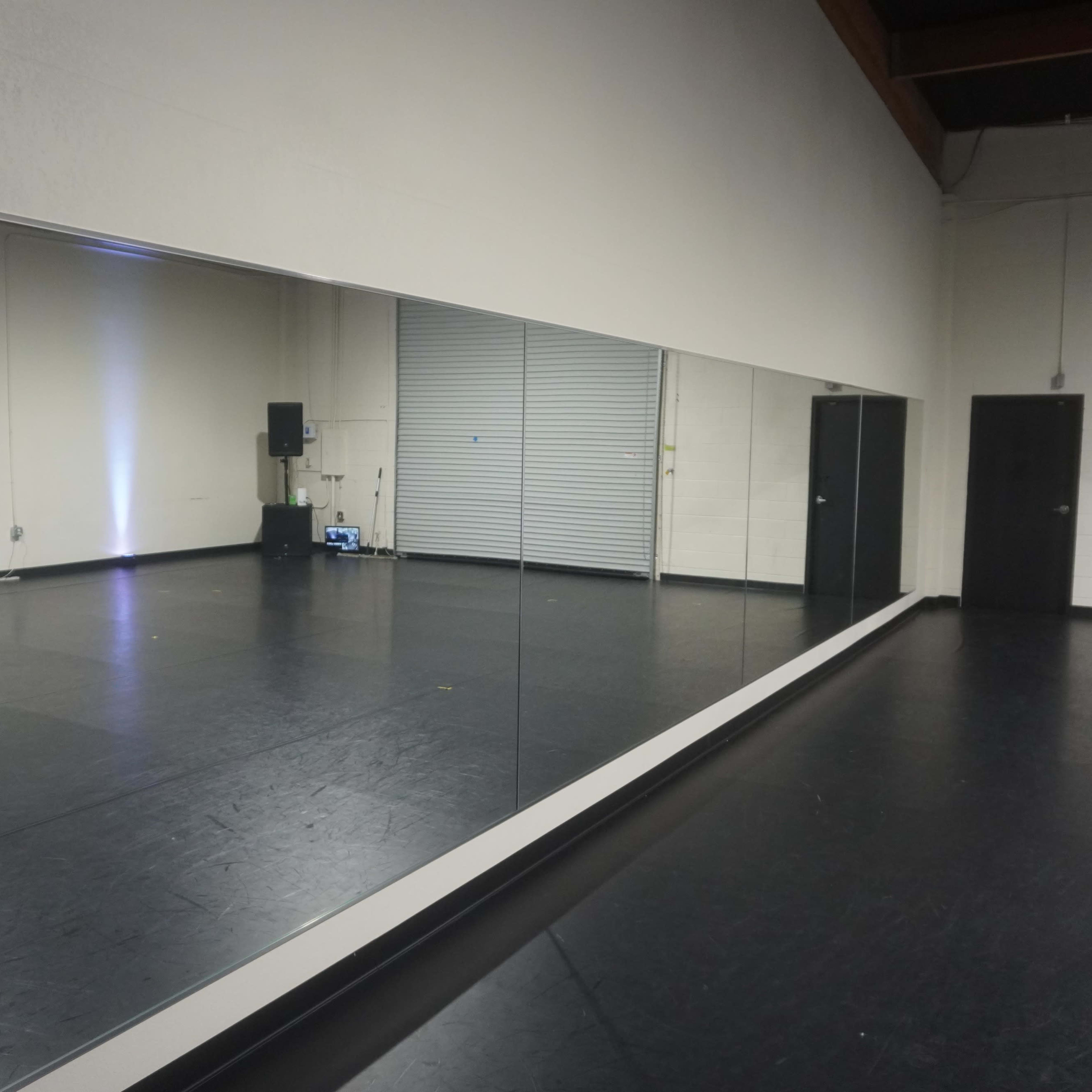 A large wall mirror reflecting a spacious, empty studio with a black floor and a closed door at one end.