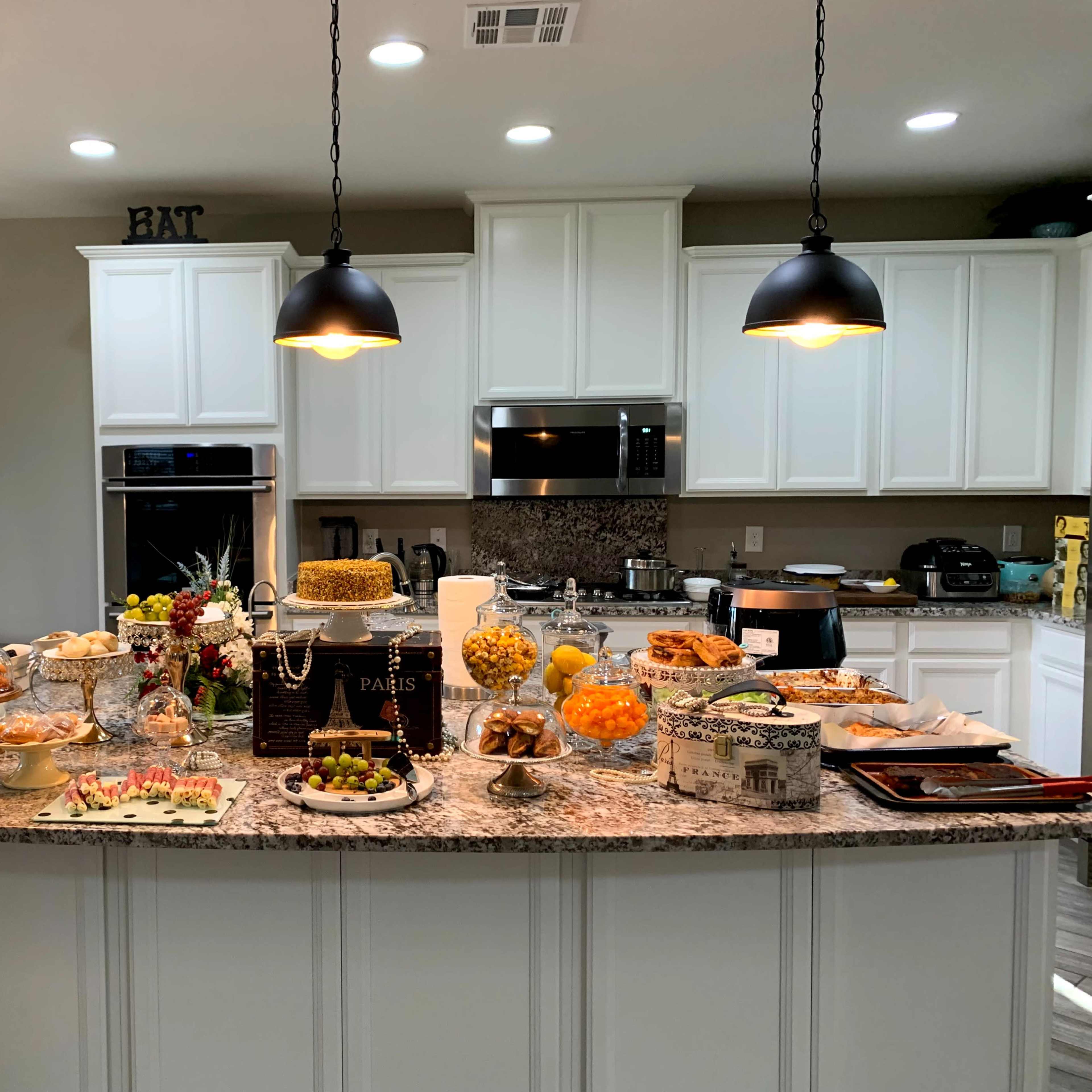 A kitchen island filled with a variety of dishes, desserts, and fruits, under hanging pendant lights.