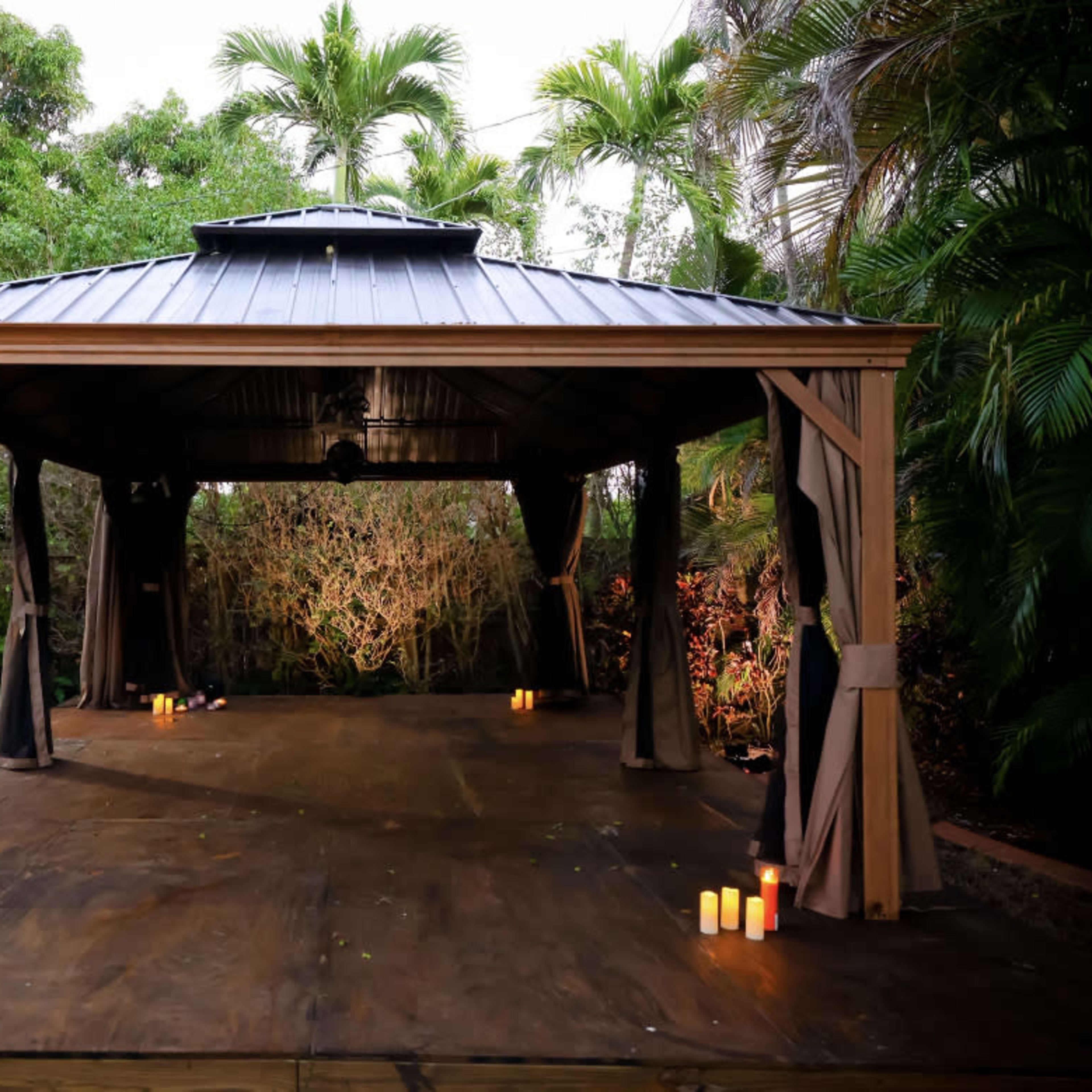 An outdoor pavilion with a dark wooden floor is surrounded by lush greenery and illuminated by candles placed on the ground.