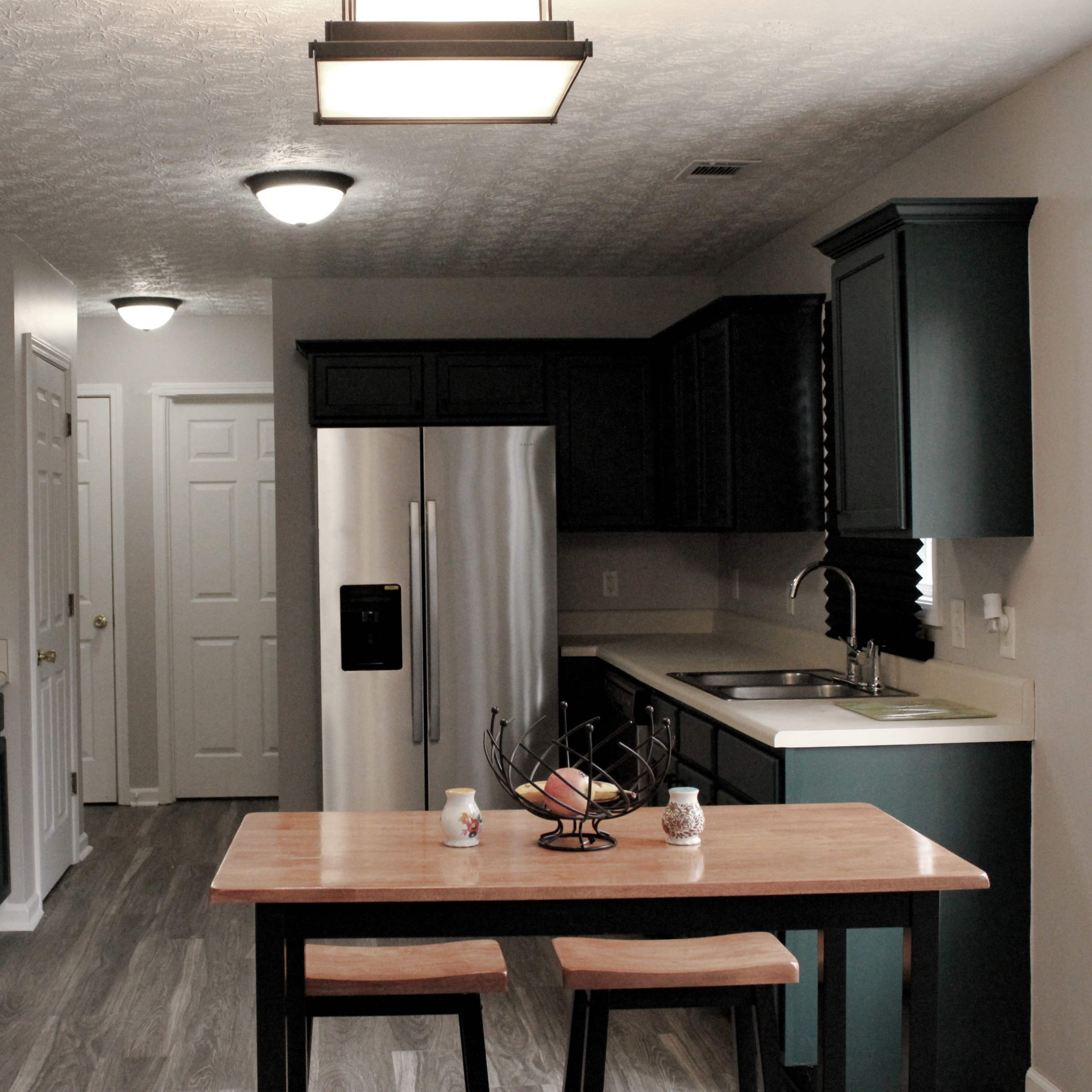 A modern kitchen featuring dark green cabinets, stainless steel appliances, and a small wooden dining table with a fruit basket.