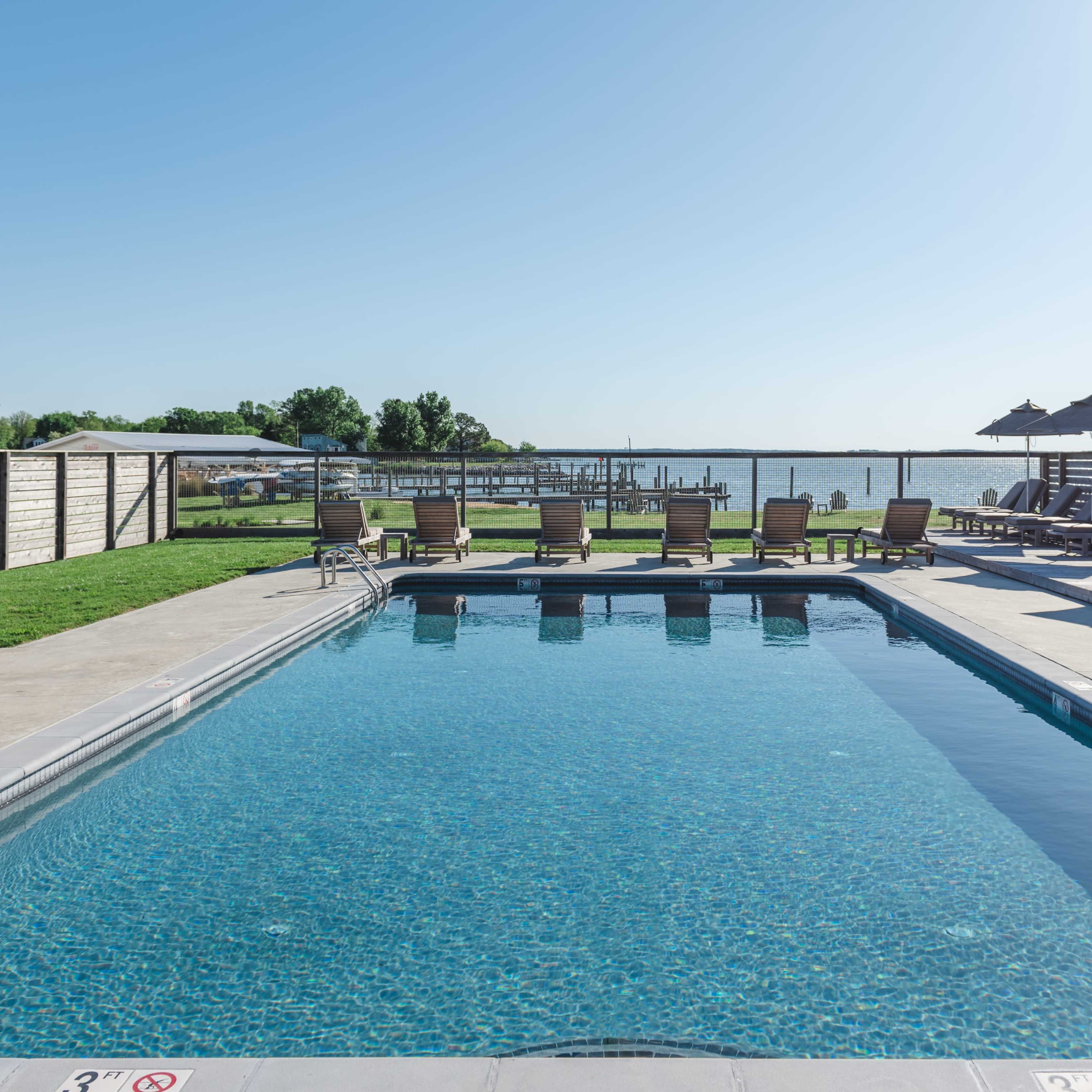A clear swimming pool surrounded by lounge chairs and umbrellas, with a view of a waterfront in the background.