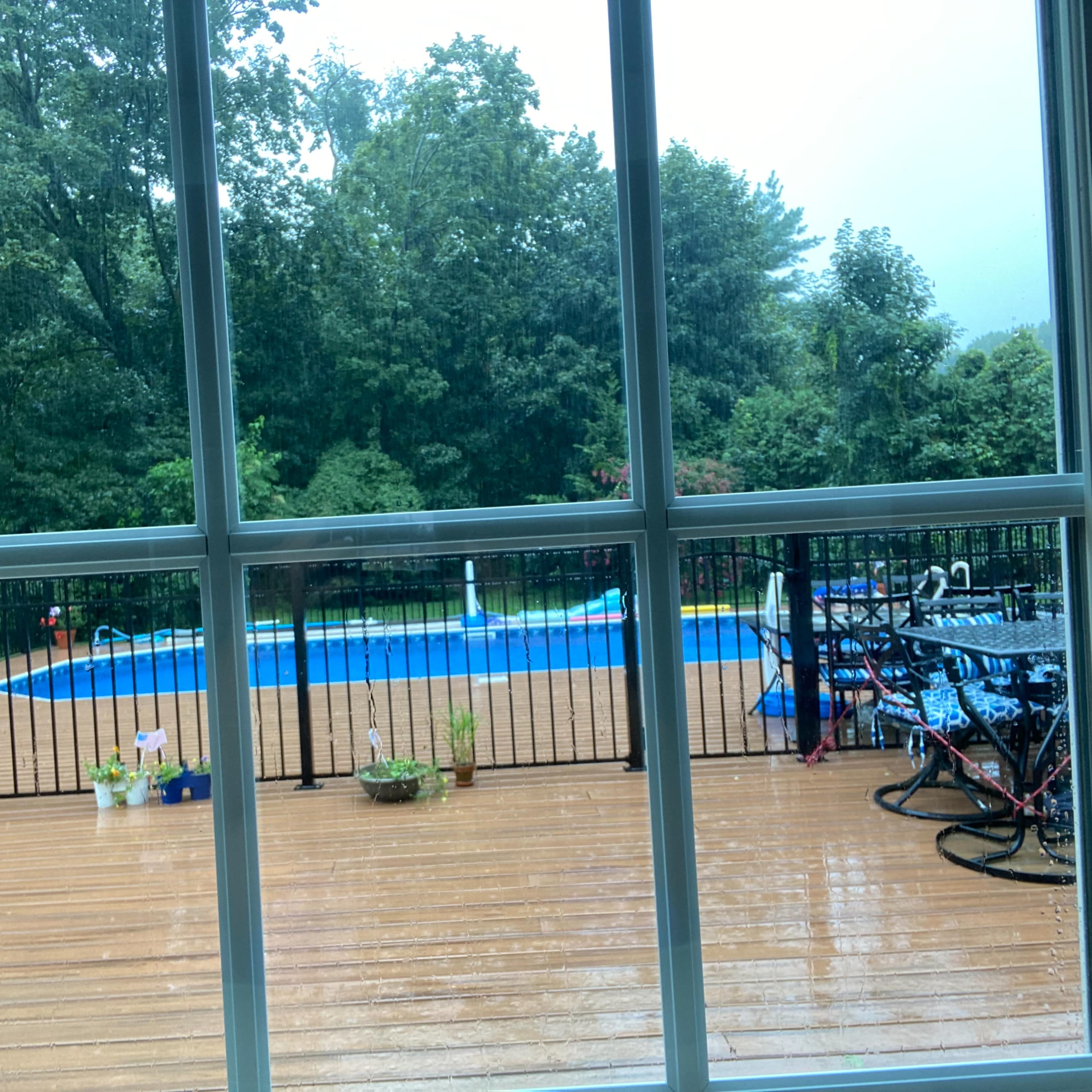 The view shows a rainy day from inside a house, looking out at a wooden deck with potted plants and a swimming pool surrounded by a black fence.