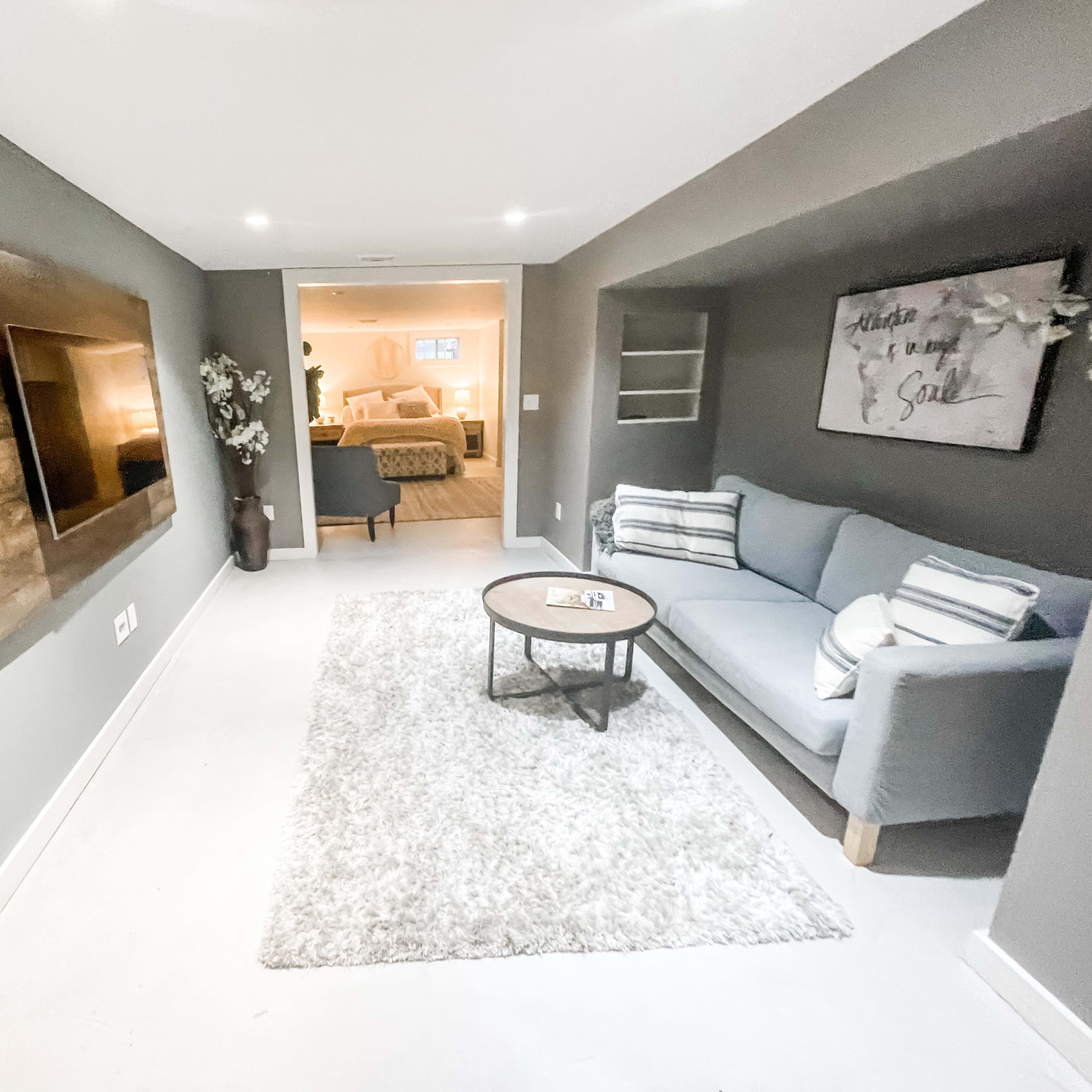 A modern basement living area features a gray sofa, a round table, and a wall-mounted TV, with light streaming in from an open doorway.