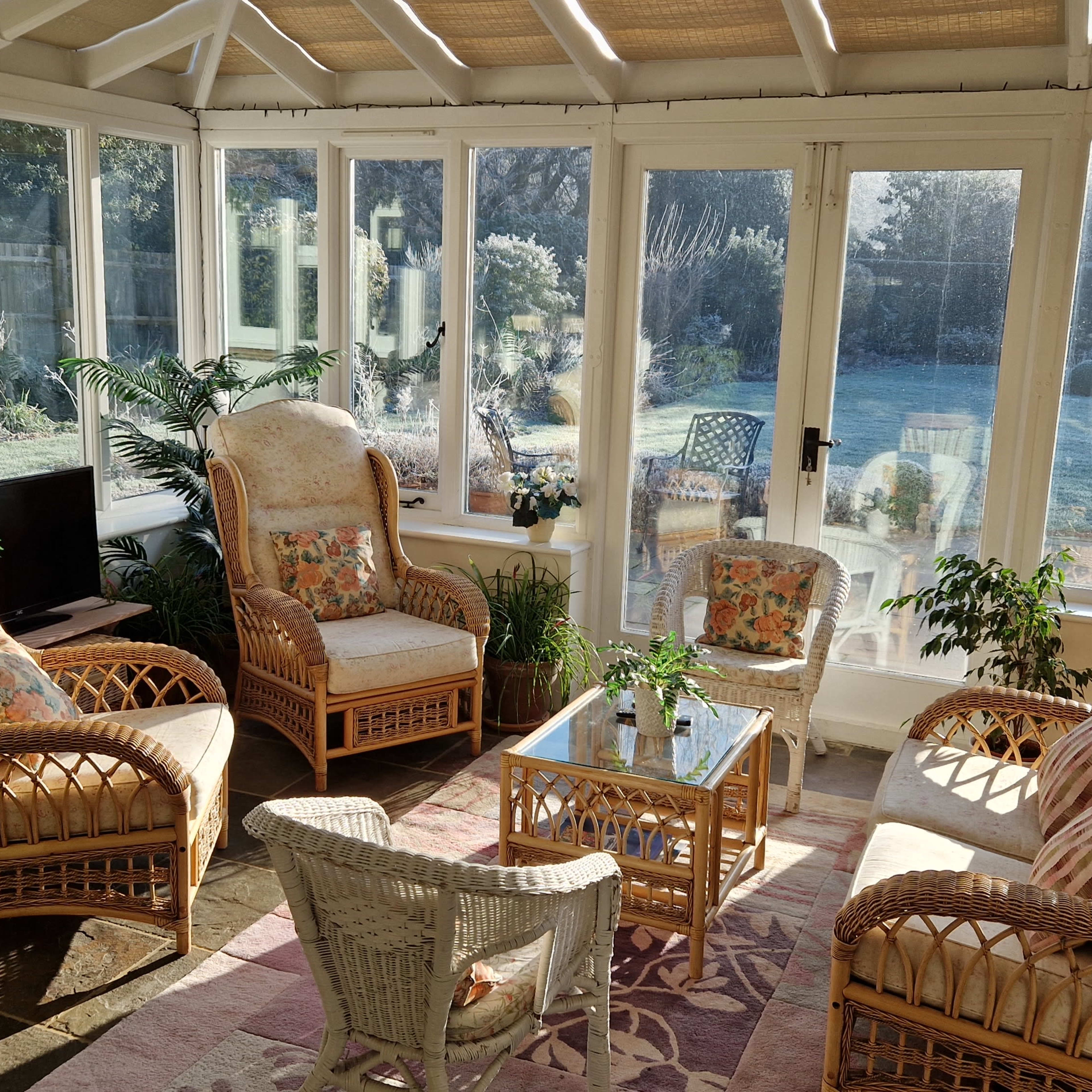 A sunlit conservatory features rattan furniture, including armchairs and a coffee table surrounded by indoor plants.