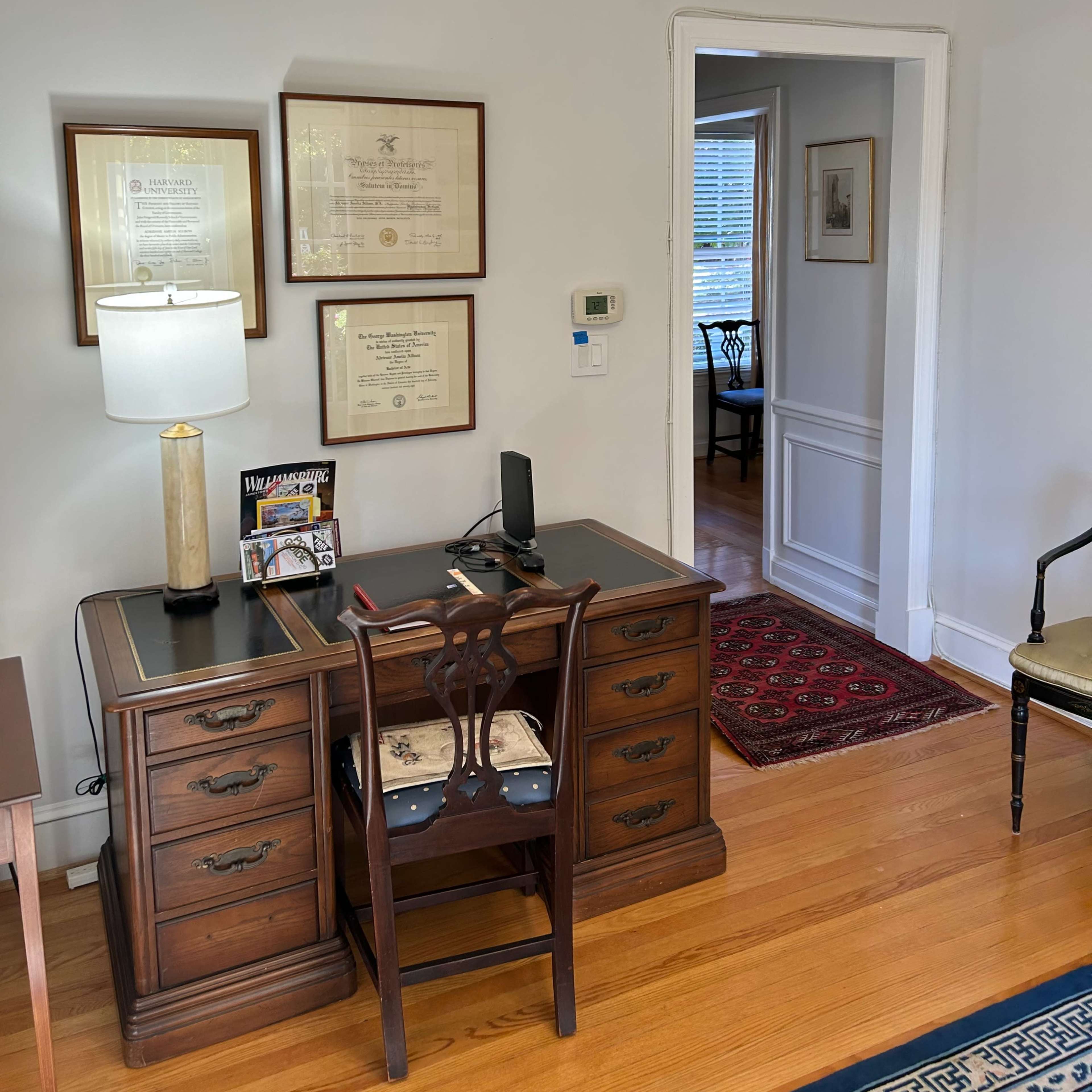 An office space features a wooden desk with a lamp, framed certificates on the wall, and a doorway leading to another room.