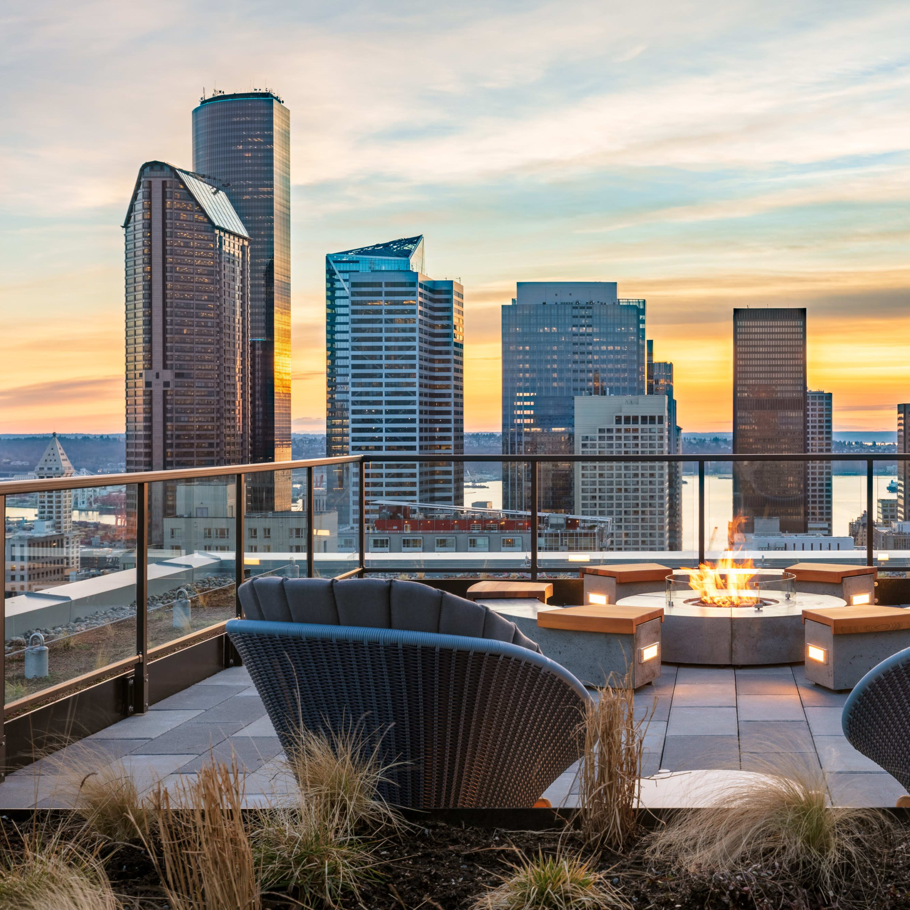 A rooftop lounge area with two chairs facing a central fire pit, overlooking a skyline of modern skyscrapers at sunset.