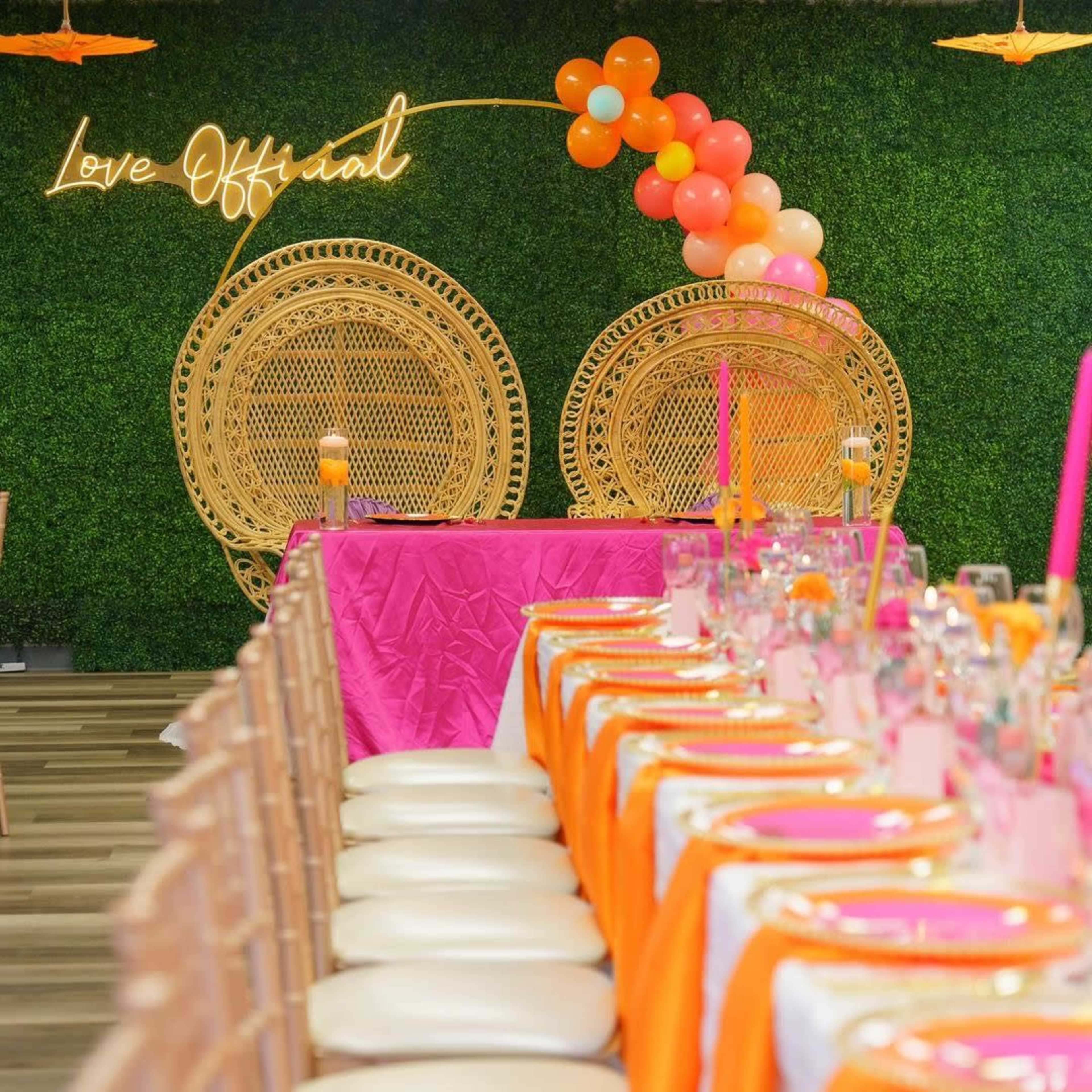 A decorated event space with a long table set with orange and pink tableware, flanked by two large wicker chairs and a neon sign that reads "Love Official."