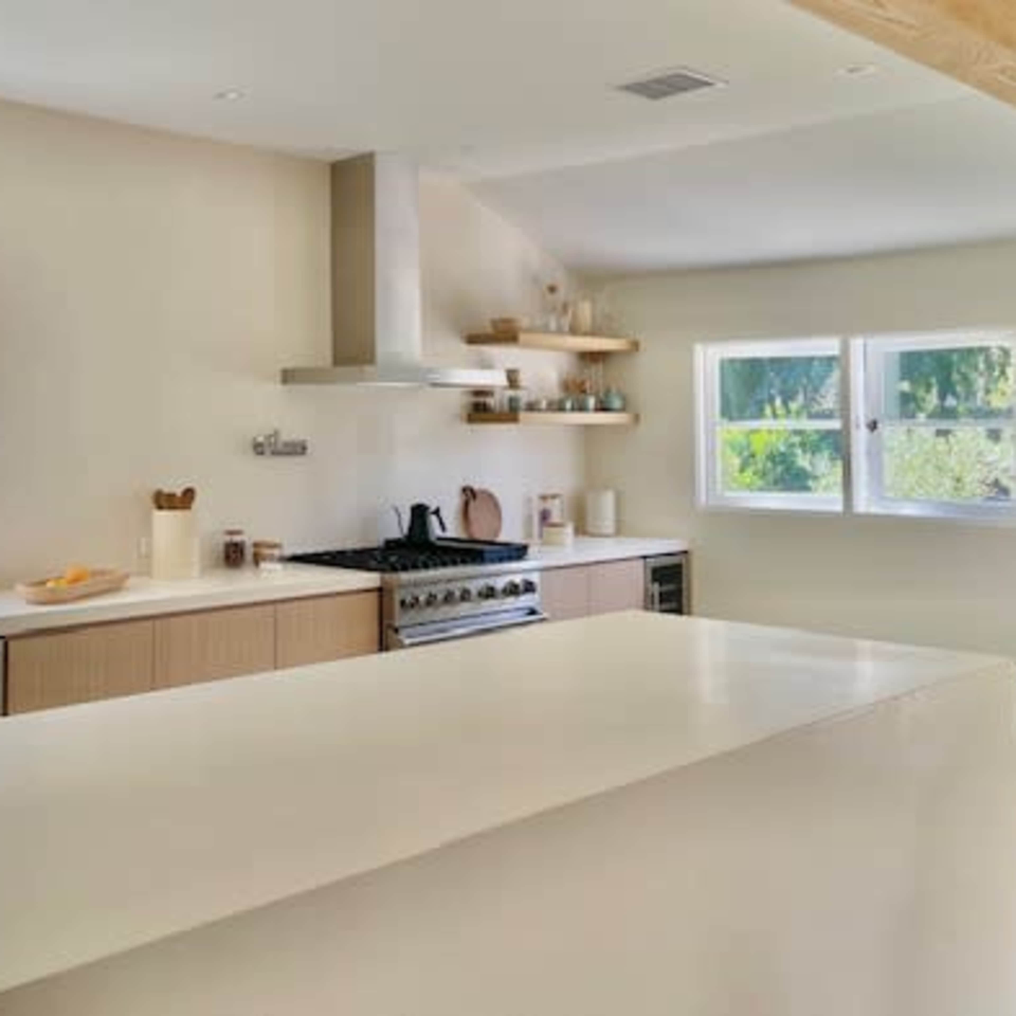A modern kitchen with a large island, light wood cabinetry, and a gas stove against a neutral-colored wall, illuminated by natural light from the windows.