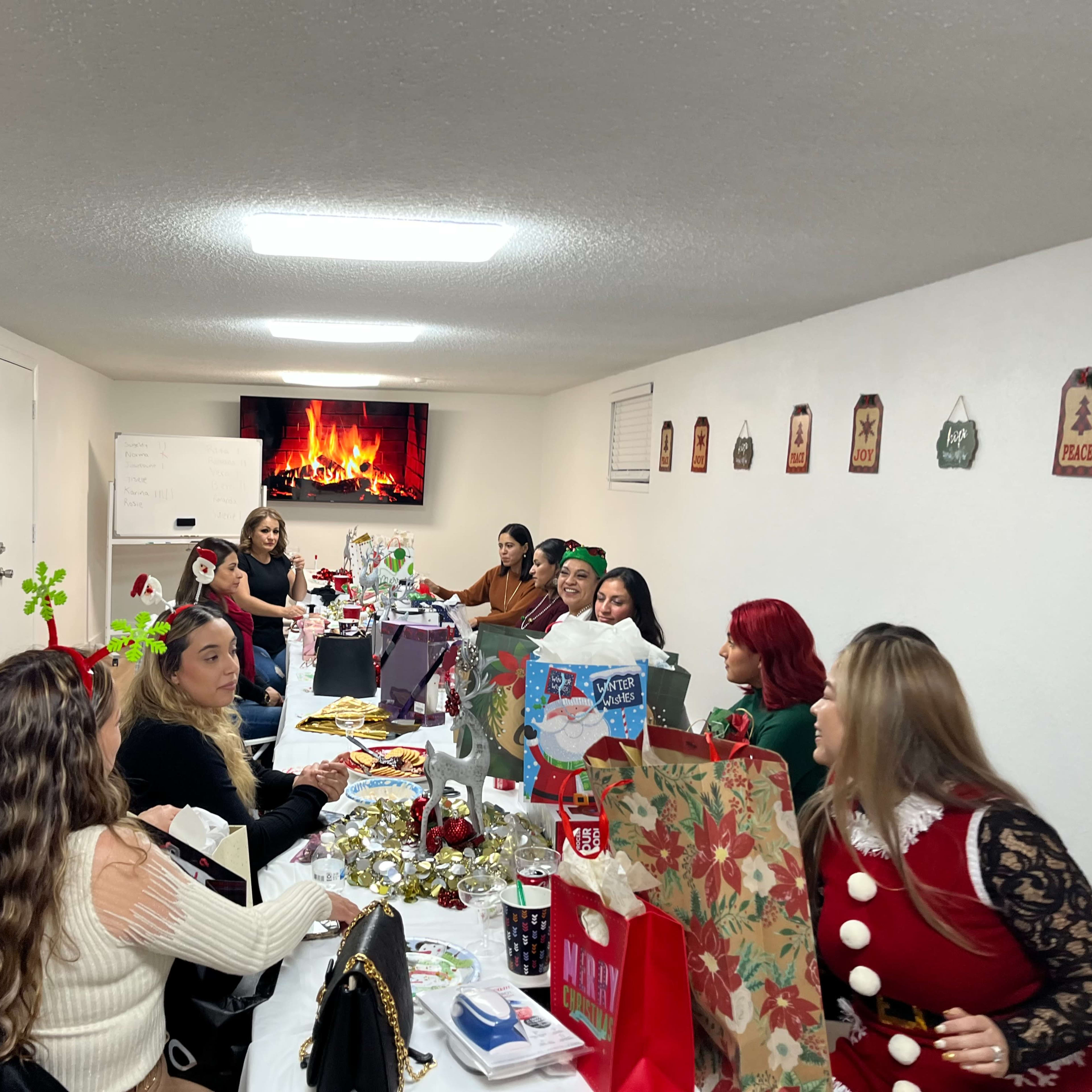 A group of people sit around a long table decorated for a holiday celebration, while a festive display of gifts and decorations is visible.