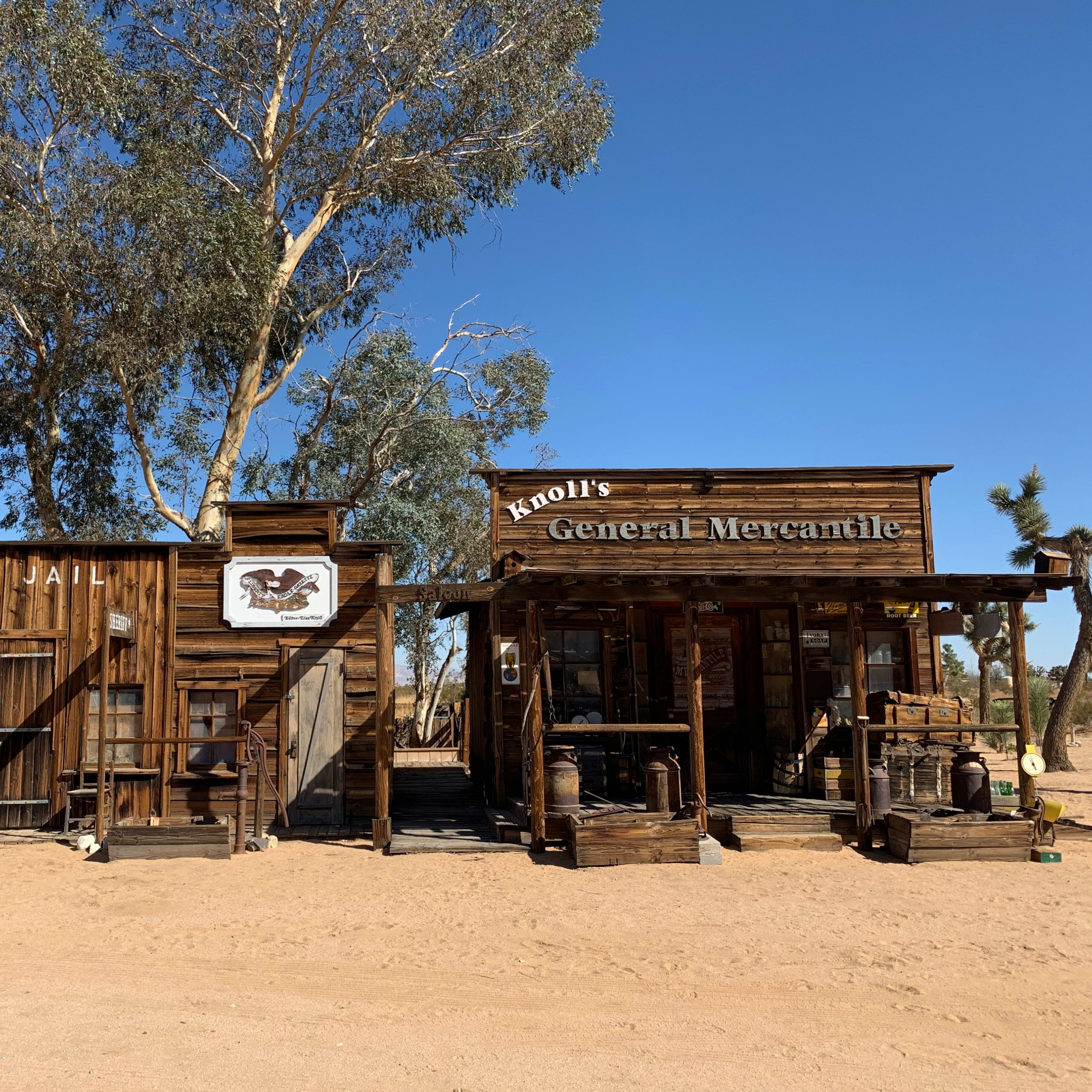 A rustic general mercantile store and jail, both made of weathered wood, set against a clear blue sky and sandy ground in a desert landscape.